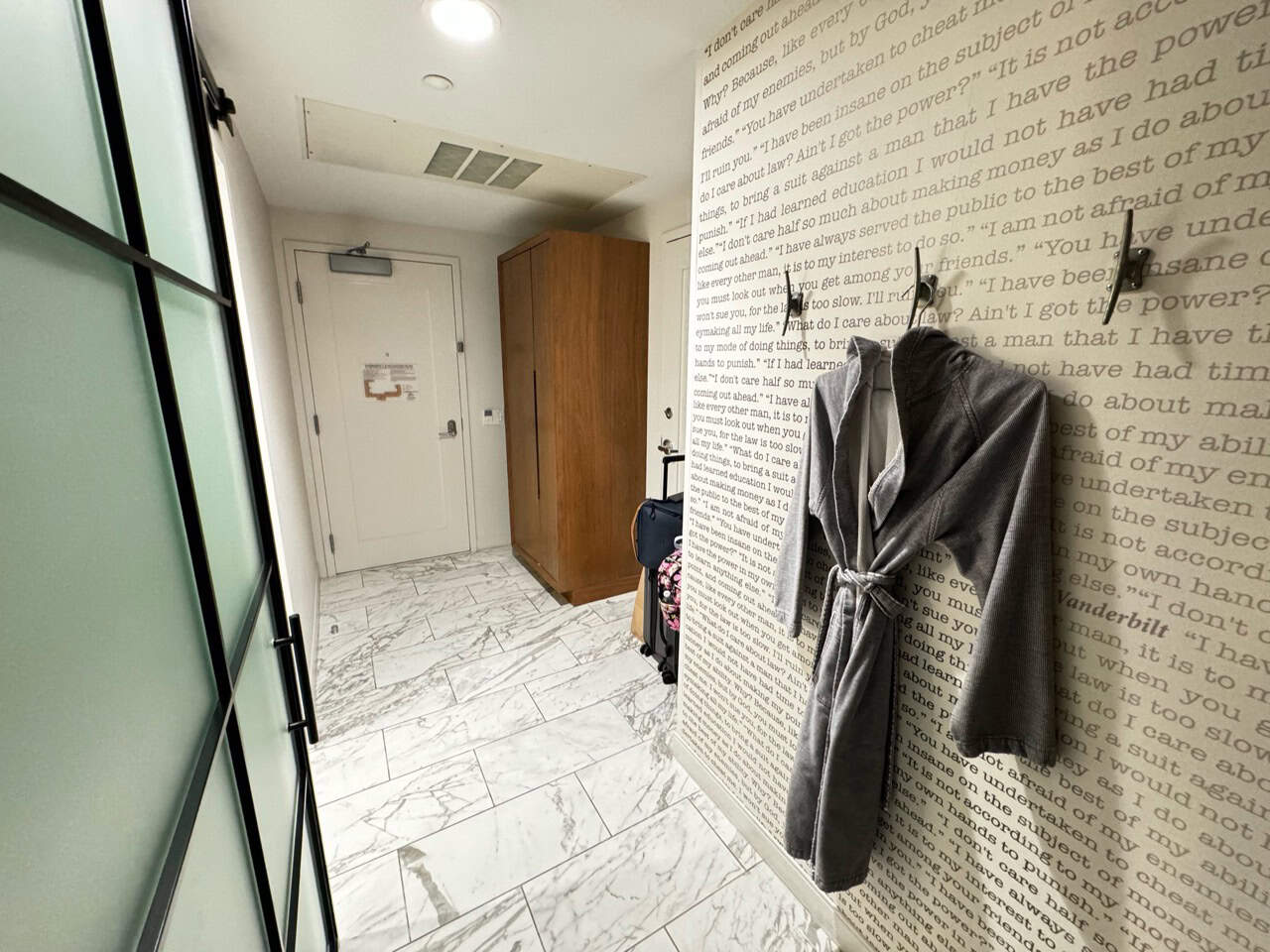 A Nashville-inspired bathroom features a pristine white marble floor, a wooden wardrobe, and wall hooks holding a gray robe. The opposite wall showcases an artistic text print design, adding a touch of Southern charm.