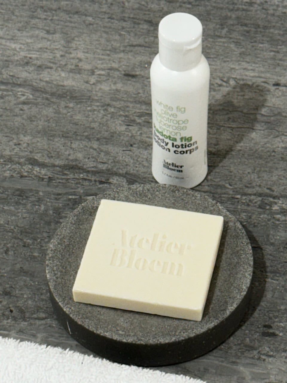 A square soap bar with "Atelier Bloom" engraving sits on a dark stone dish, evoking the rustic charm of Nashville. A small white bottle of body lotion labeled "White Fig Olive" is positioned behind it on a gray countertop.