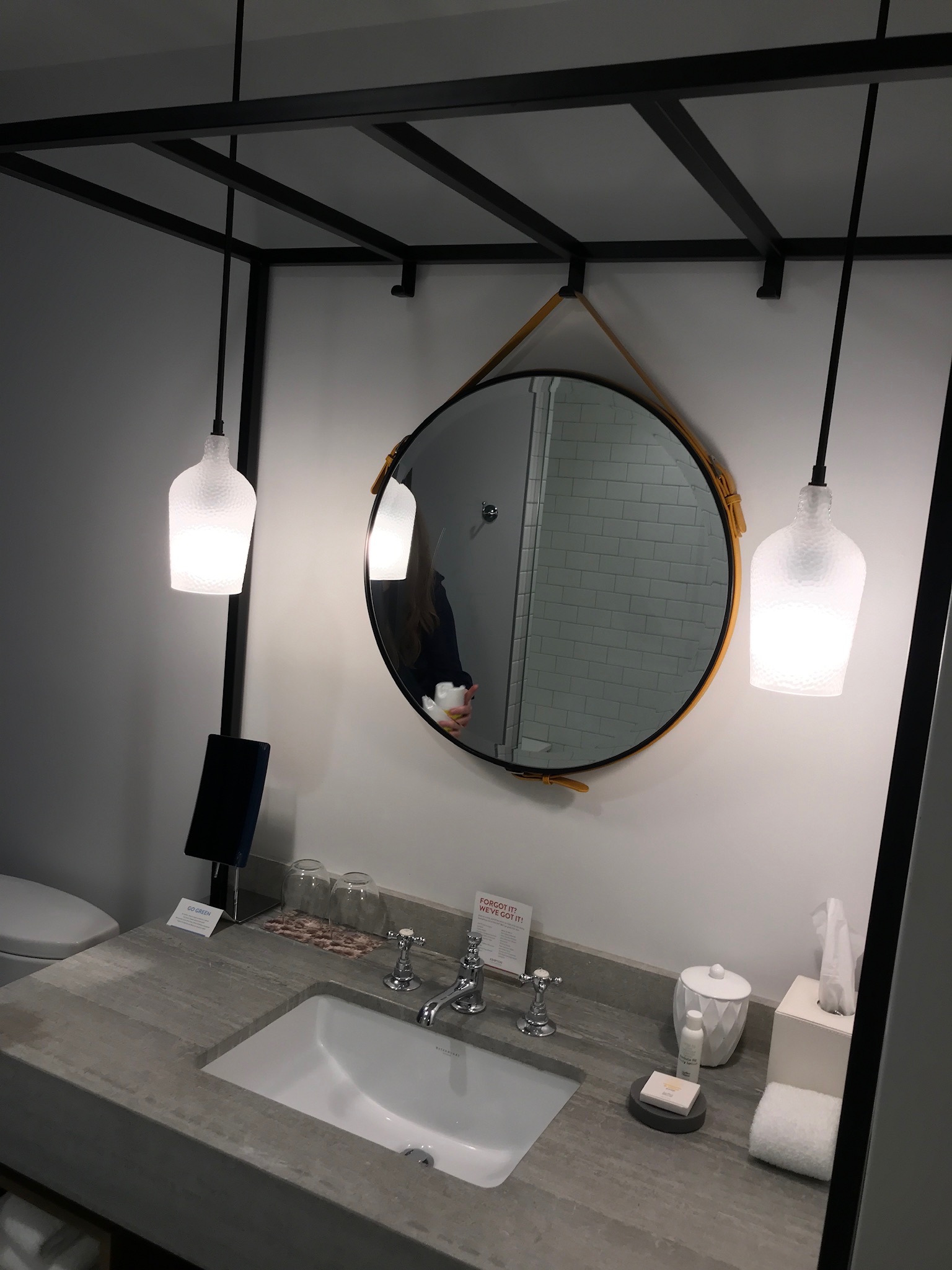 Modern bathroom vanity with a round mirror, pendant lights, a rectangular sink, tissue box, soap, and amenities on a concrete countertop. White tiled shower reflected in the mirror.