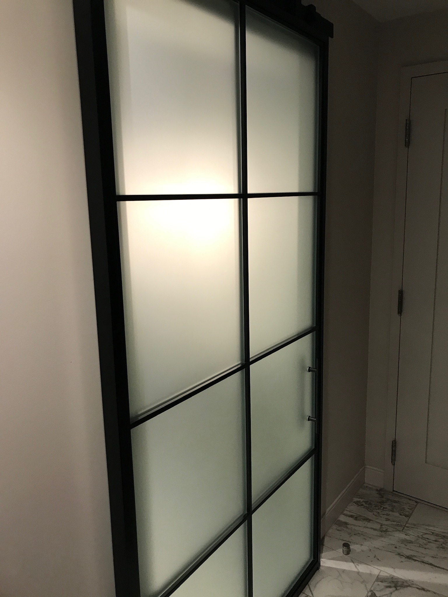 A frosted glass sliding door with a black metal frame and grid pattern, set against a beige wall and marble floor indoors.