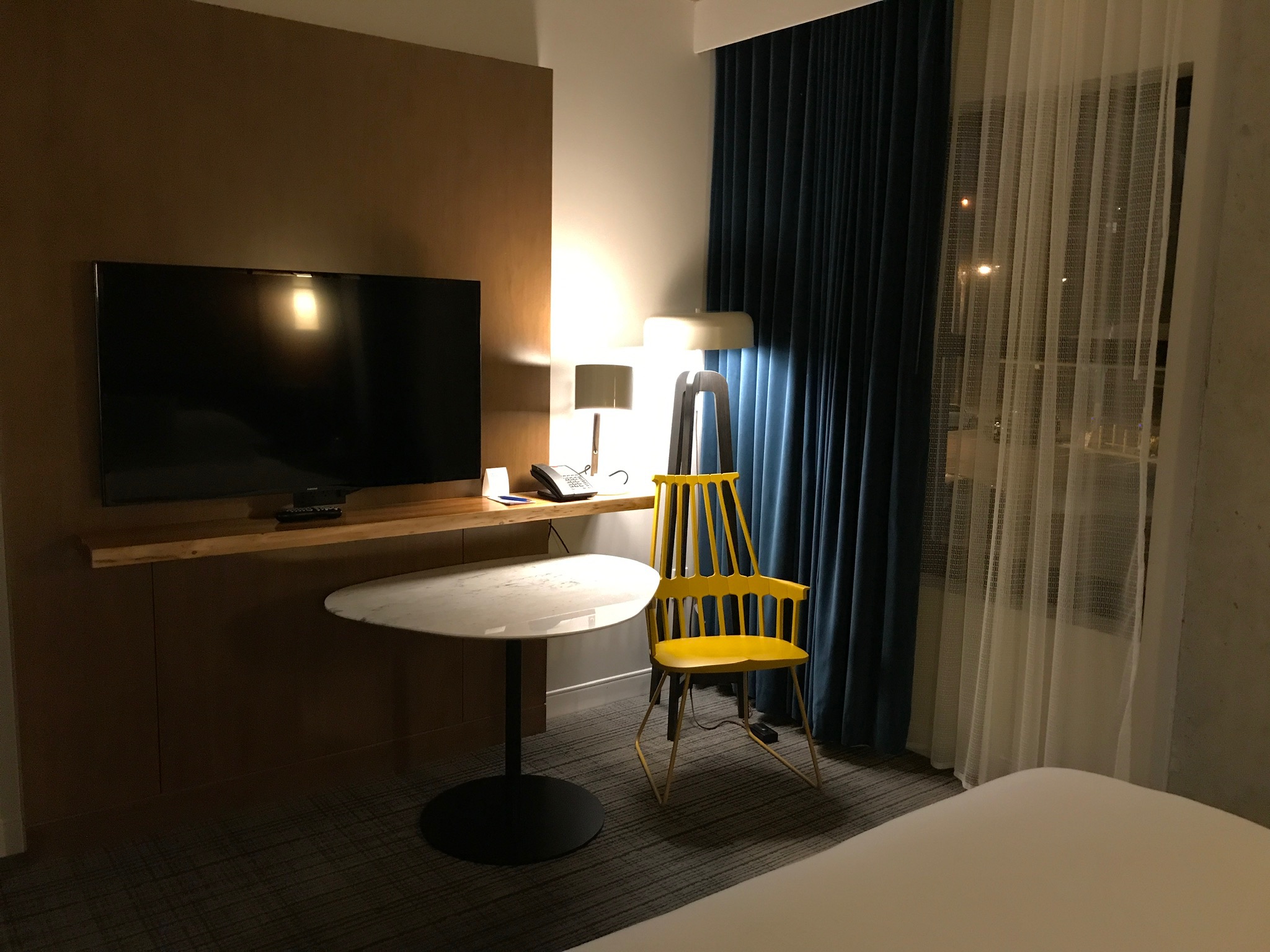 A hotel room with a flat-screen TV on a wooden panel, a round table, a yellow chair, a lamp, dark curtains, and white sheer drapes by a window at night.