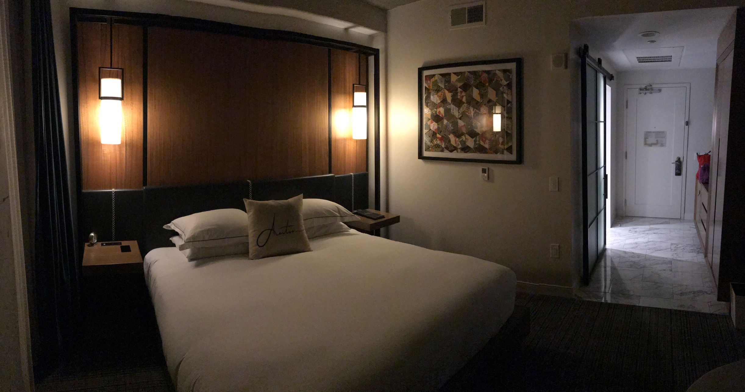A hotel room with a neatly made bed, two wall lamps, framed artwork on the wall, and an open entryway with marble flooring visible.