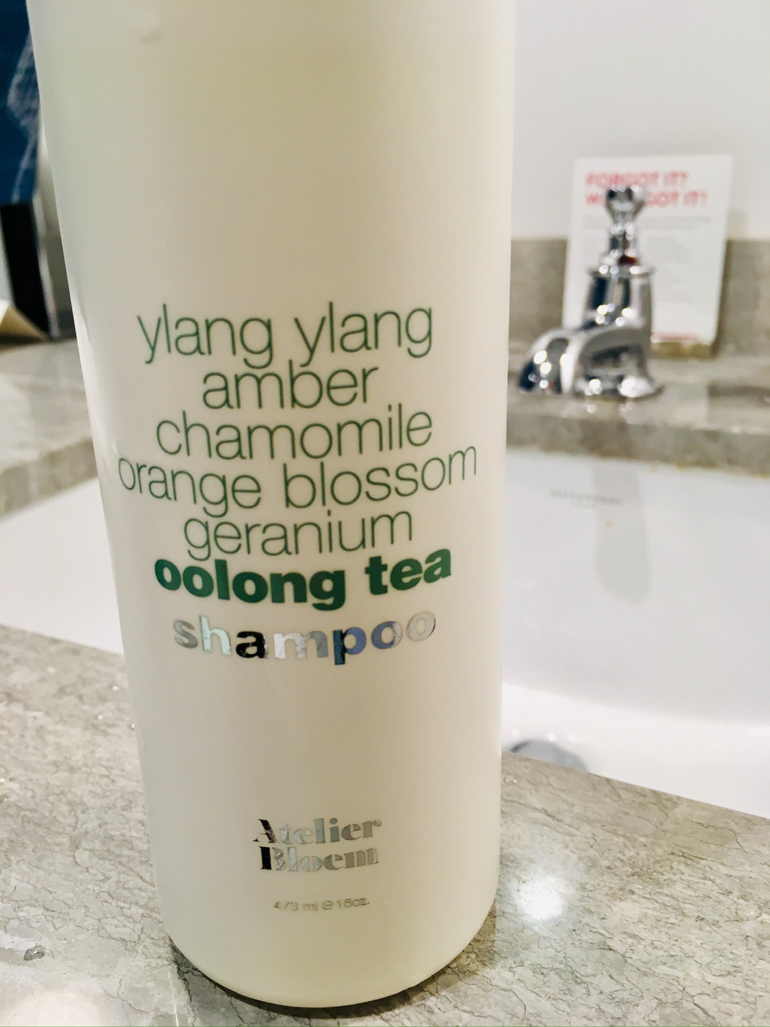 A white bottle of Atelier Bloem shampoo with ingredients such as ylang ylang, amber, chamomile, orange blossom, geranium, and oolong tea listed on the label.