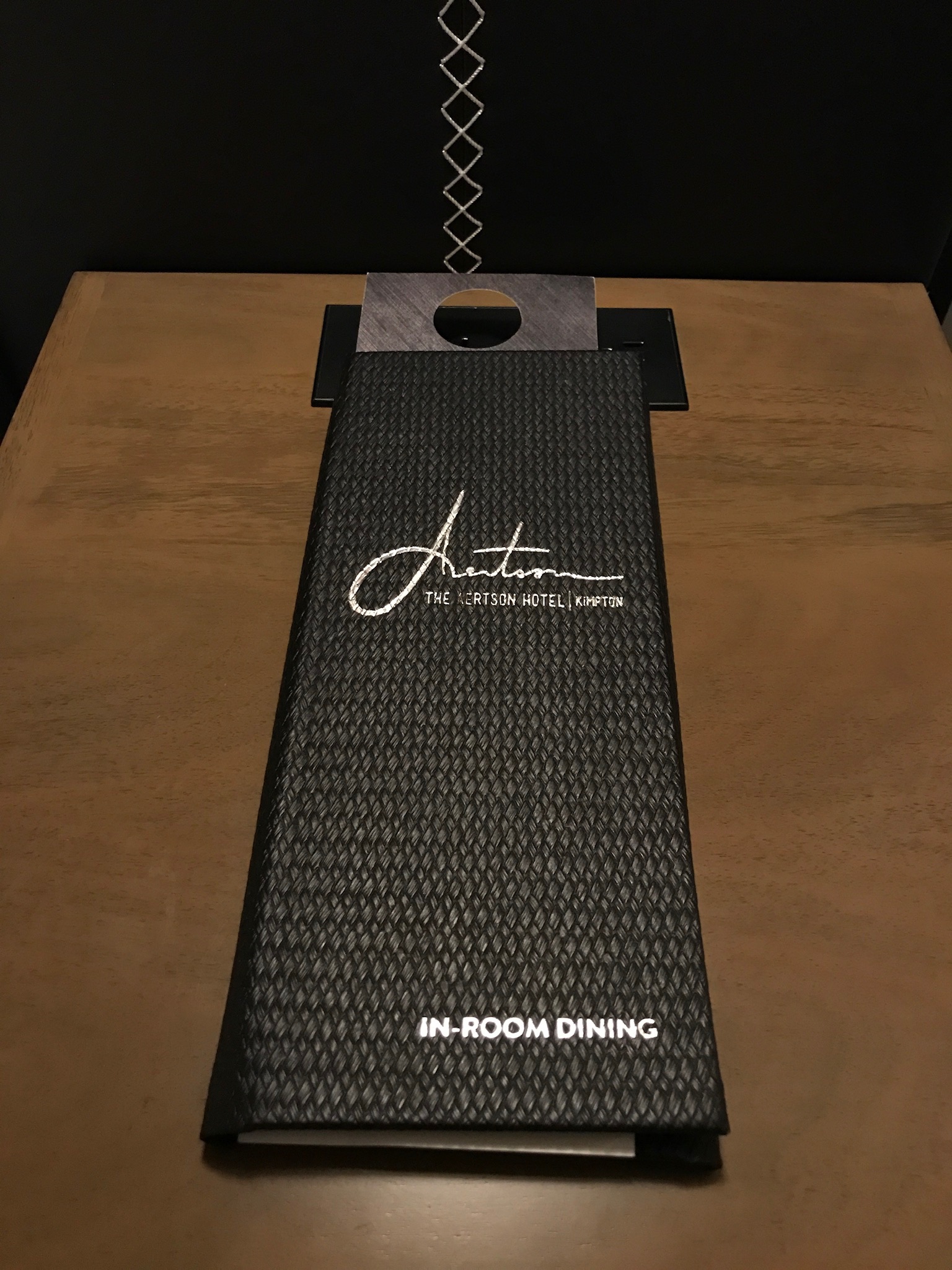 A black textured folder labeled "IN-ROOM DINING" from The Artisan Hotel, Kempton, placed on a wooden surface under a hanging pendant light.