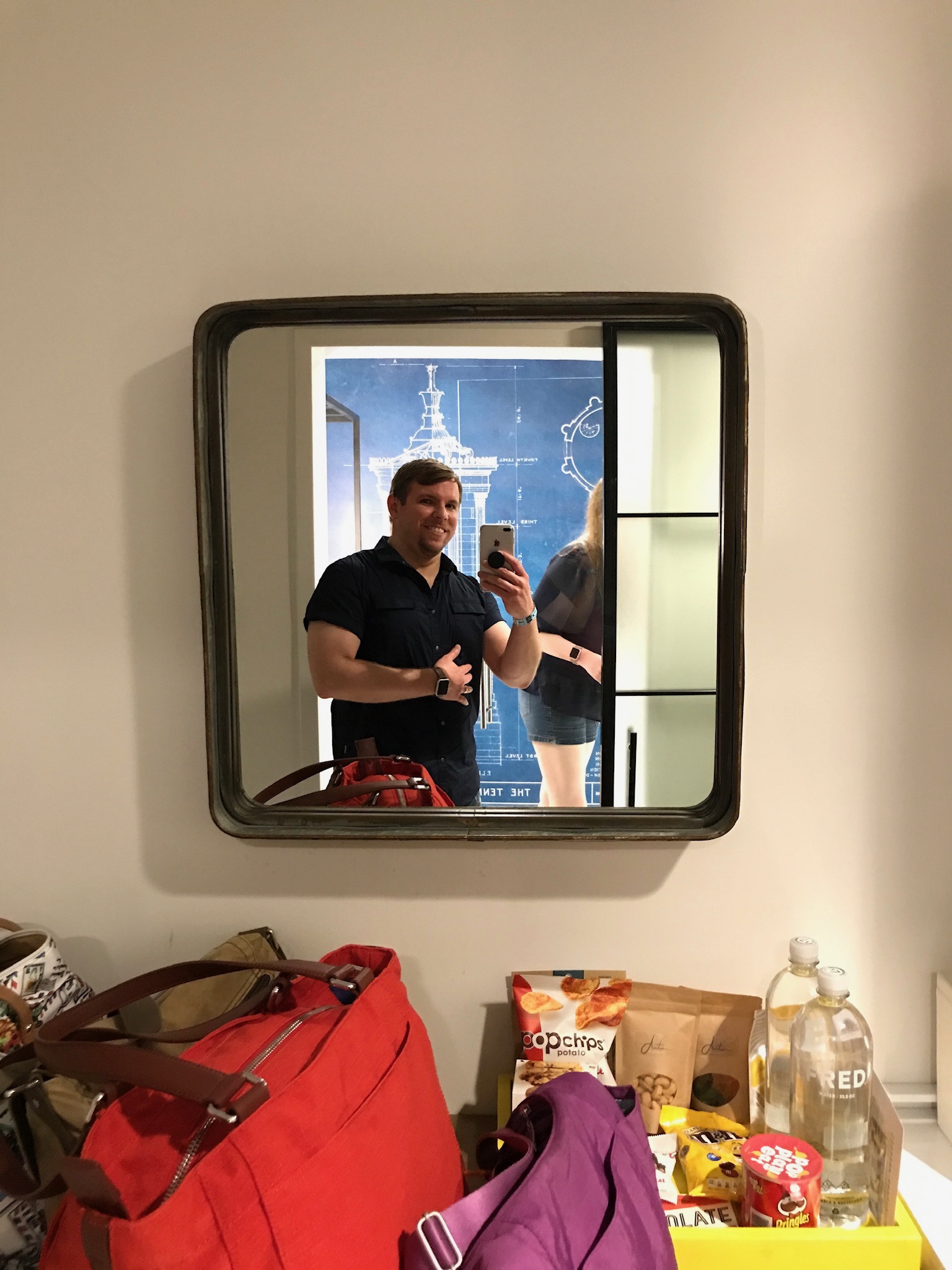 A man takes a selfie in a wall mirror; a woman stands partially out of frame. Various bags, snacks, and drinks are on a table below the mirror.