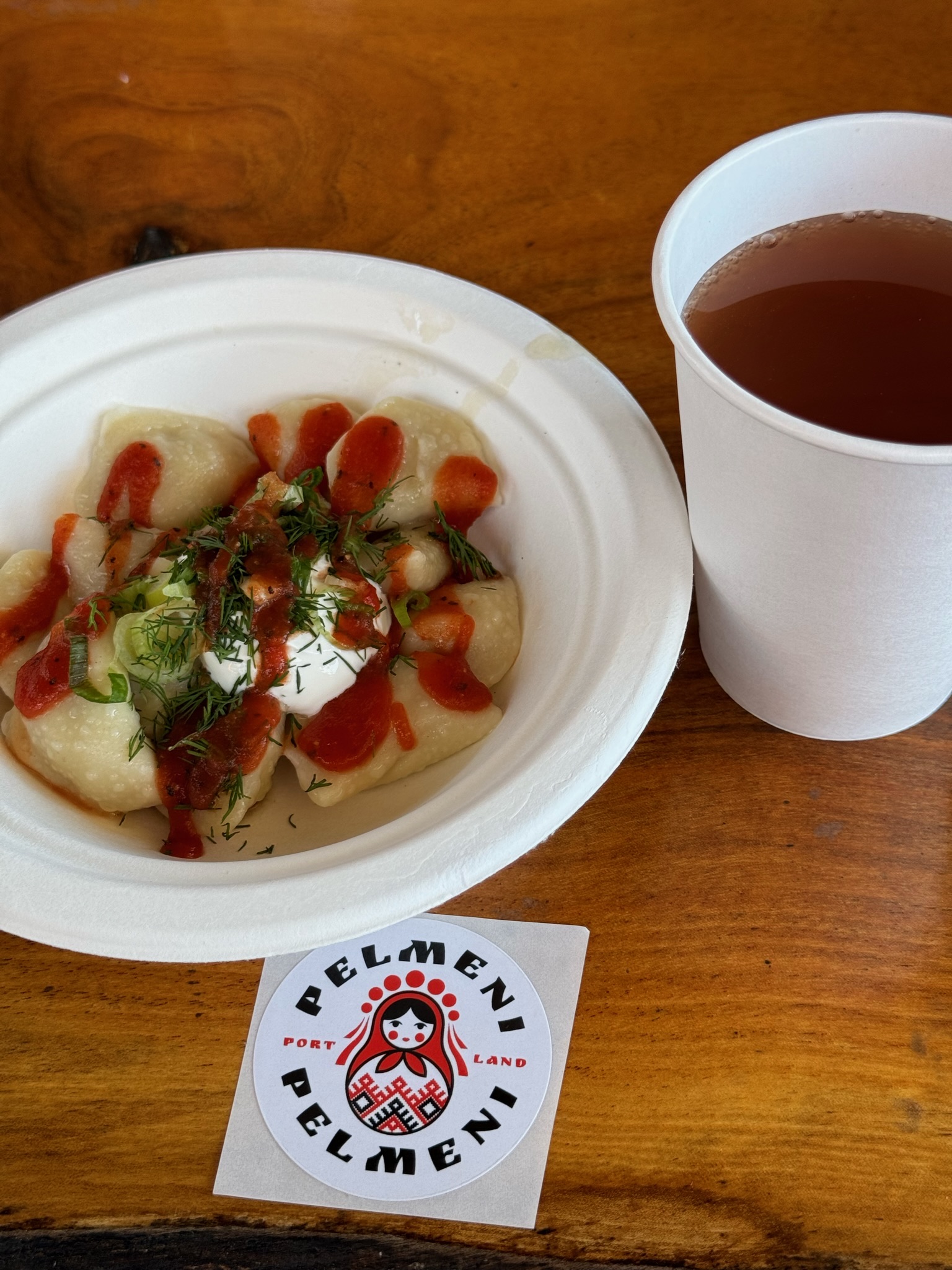 A paper plate of dumplings topped with sour cream, dill, green onions, and red sauce next to a paper cup of dark drink and a Pelmen Pelmen logo sticker on a wooden table.