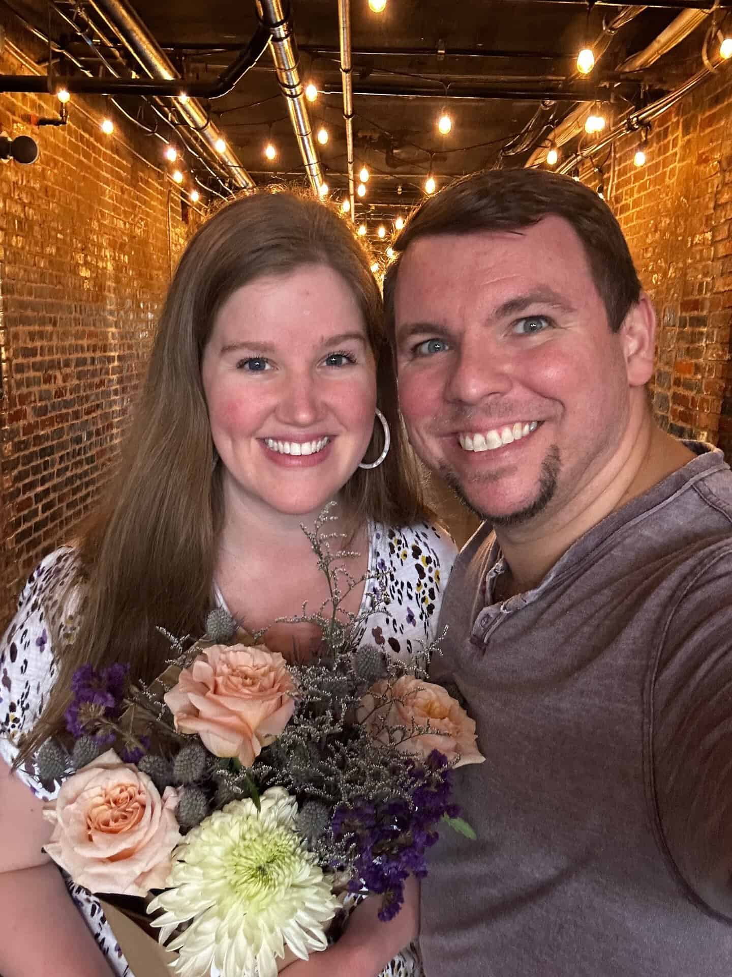 In a warmly lit hallway with exposed brick walls, a smiling woman holding a bouquet stands next to a grinning man, capturing the charming essence of Nashville.