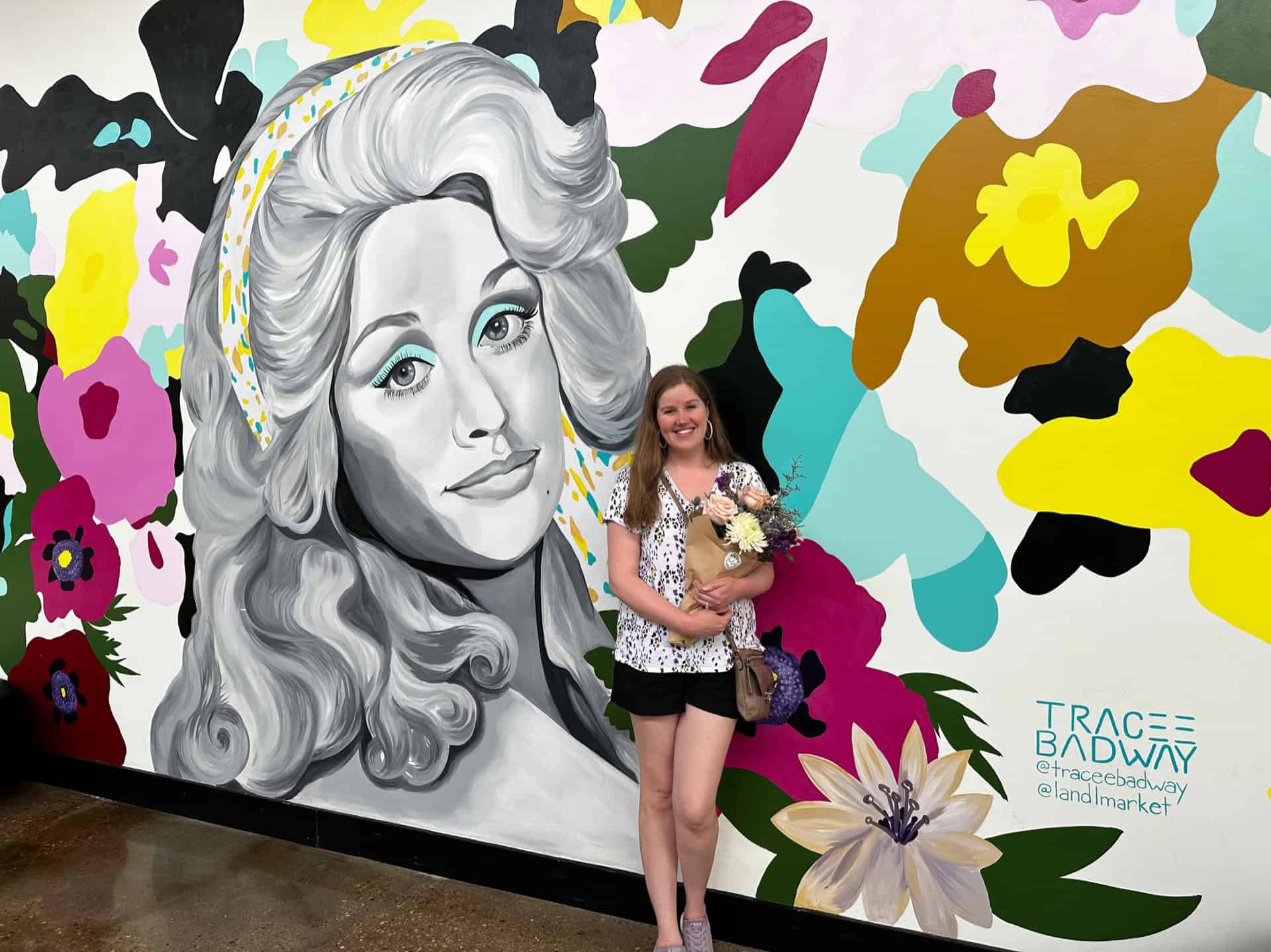 In Nashville, a woman holding flowers stands before a vibrant mural showcasing a large monochrome portrait of a woman amidst a floral backdrop.