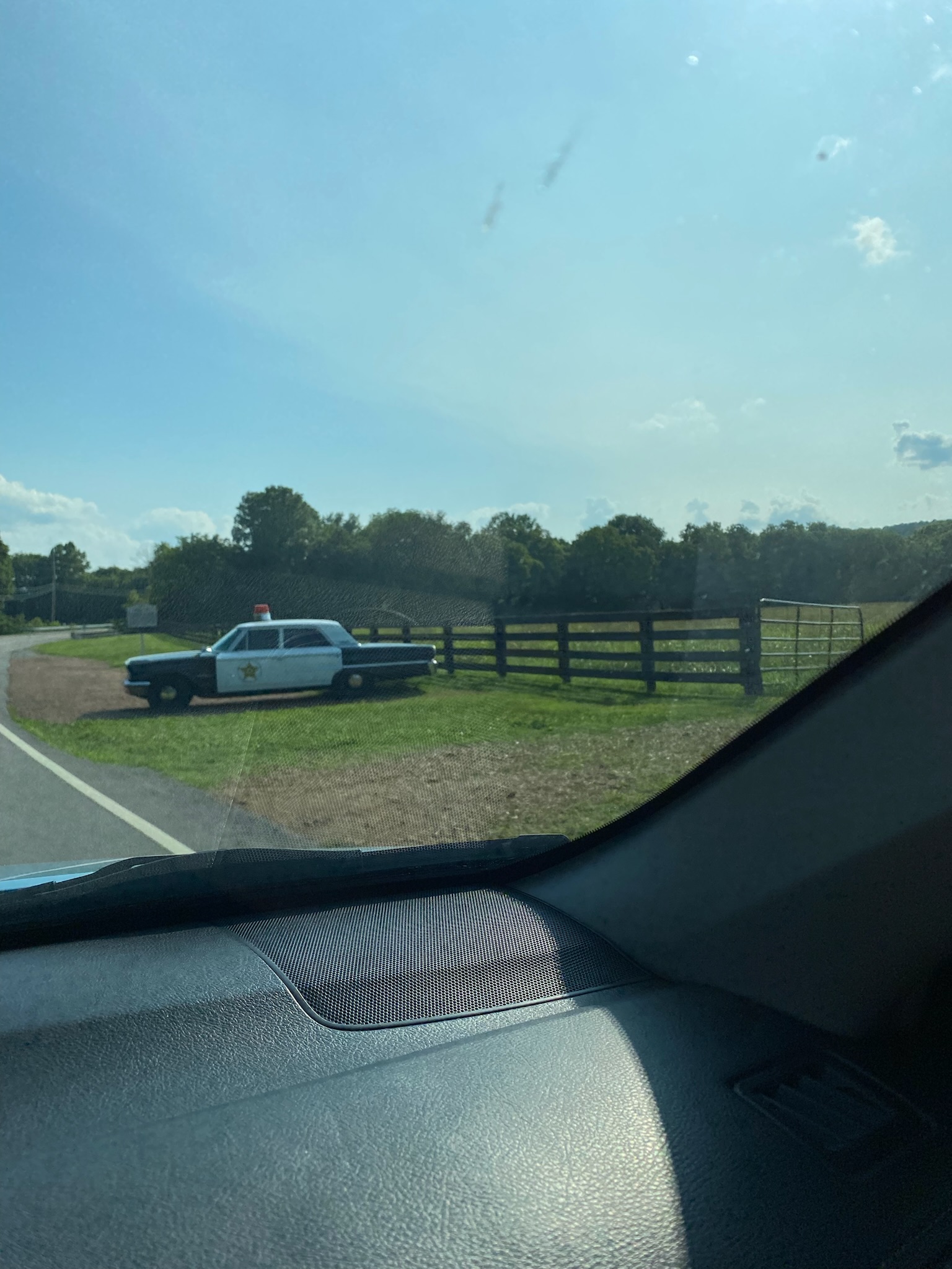 A vintage police car with a roof siren is parked on grass beside a fenced field, viewed from inside another vehicle on a sunny day.