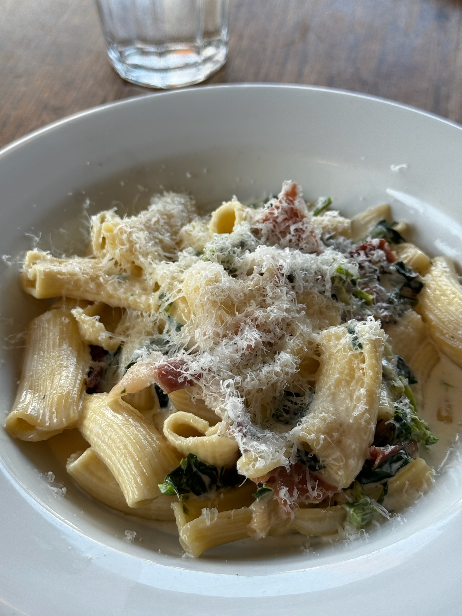 A white bowl of rigatoni pasta with greens and bacon, topped with grated cheese. A glass of water is in the background on a wooden table.