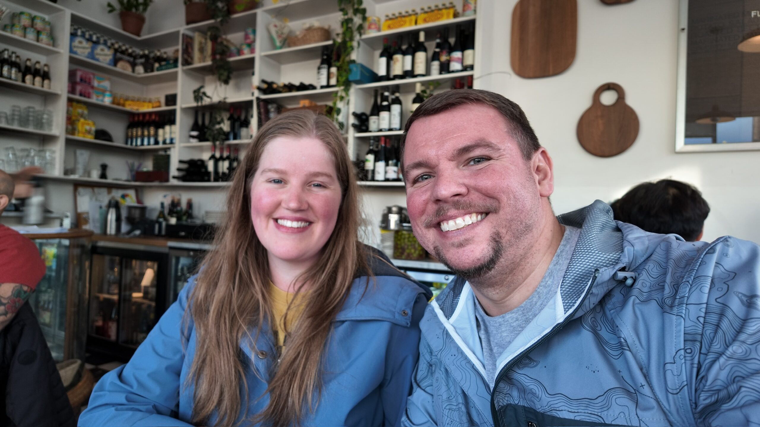 Two people wearing blue jackets smile at the camera while sitting in a cafe with shelves of bottles, jars, and plants in the background.