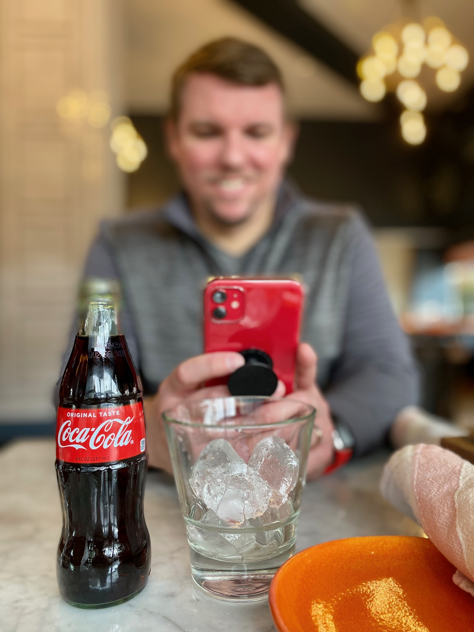 A man sits at a table holding a red smartphone. In the foreground, there is a glass bottle of Coca-Cola and a glass with ice cubes.