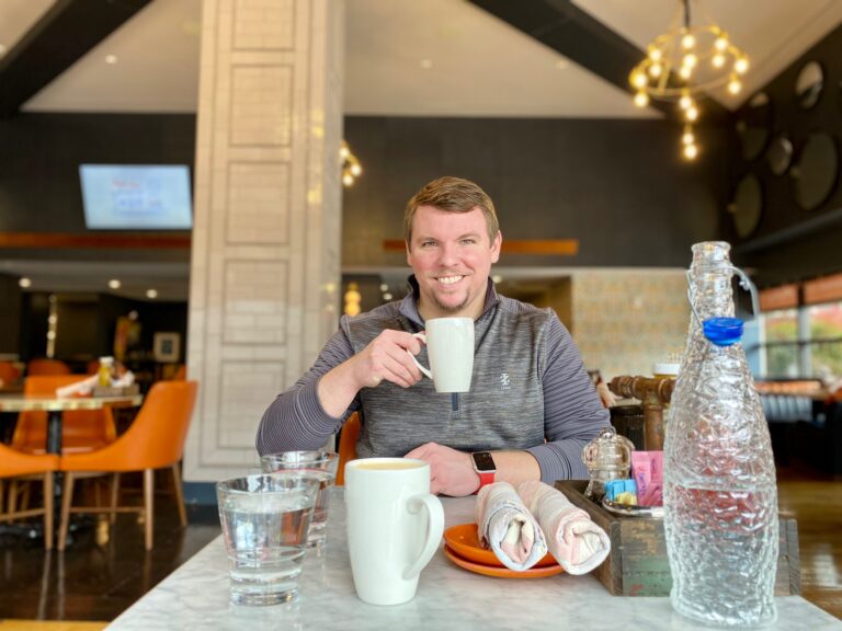 A man sits at a restaurant table holding a mug and smiling, with two glasses of water, a bottle, napkins, and another mug in front of him.