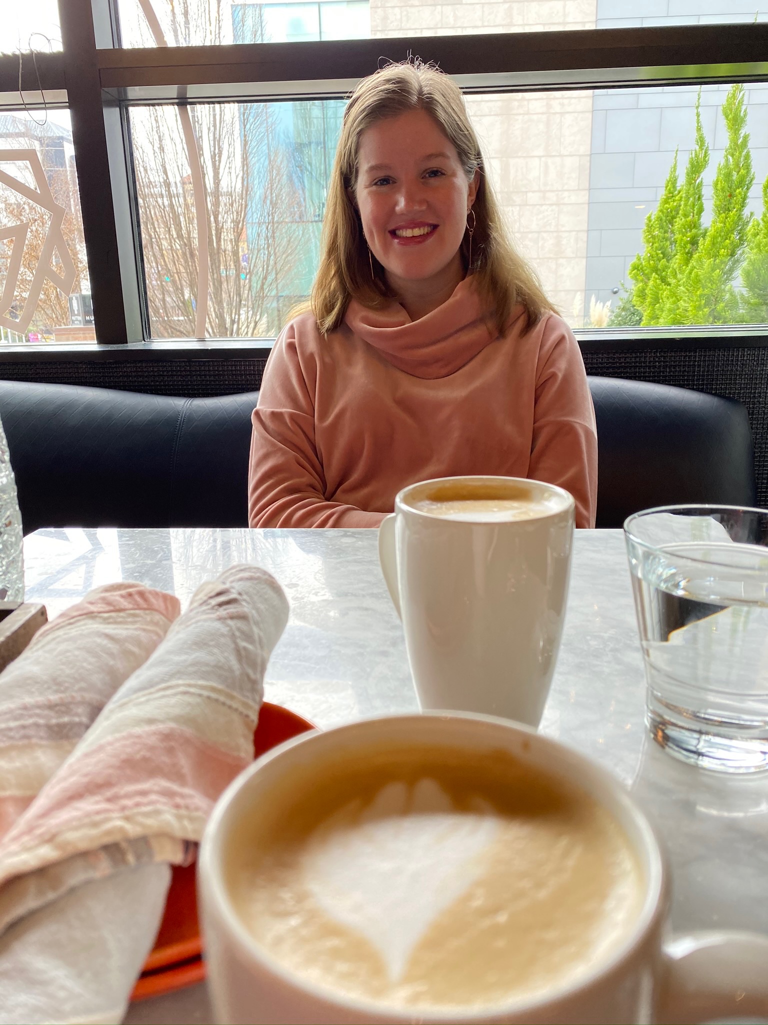 A woman sits at a table in a cafe with two cups of coffee, a glass of water, and a folded napkin visible in the foreground.