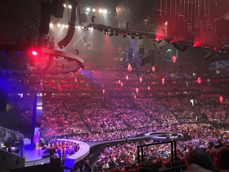 A large indoor arena filled with people, featuring a circular stage with red and purple lights, and suspended lighting equipment overhead.