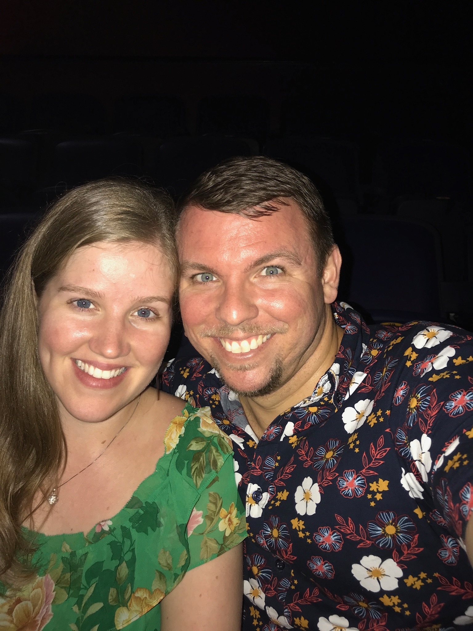A woman and a man smiling and posing for a selfie indoors. The woman is wearing a green floral top, and the man is wearing a dark shirt with a bright floral pattern.