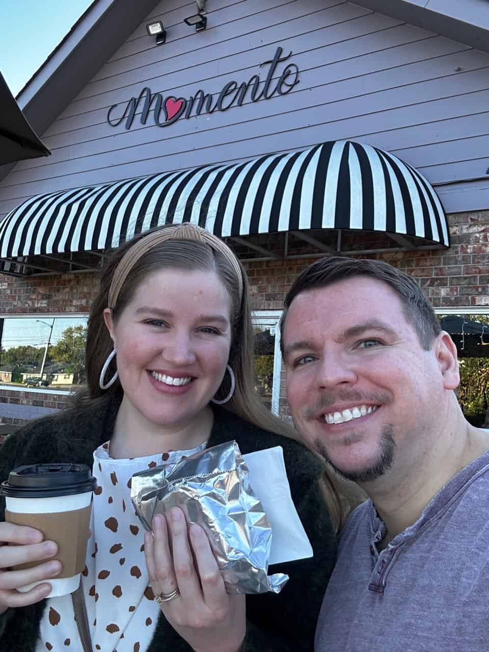 Two people smiling outside a Nashville café with a striped awning, one holding a coffee cup and a wrapped sandwich.