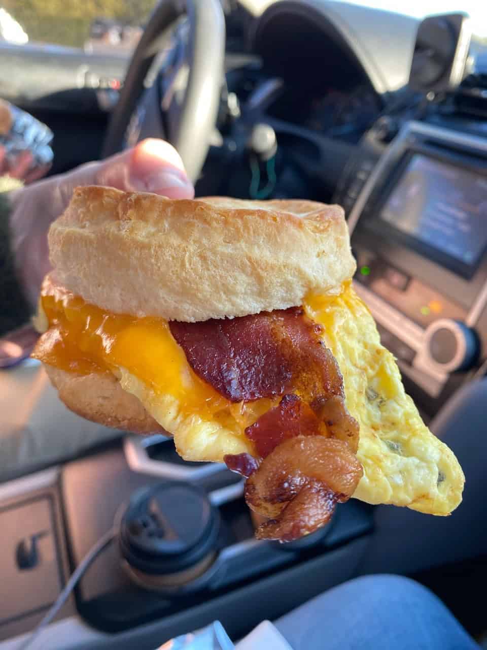 A hand holding a hearty Nashville breakfast sandwich with egg, bacon, and cheese in a car. Steering wheel and car controls are visible in the background, capturing an on-the-go breakfast moment.
