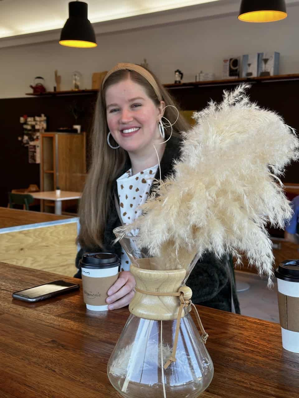 In a cozy Nashville café, a woman with long hair sits at the table, smiling as she cradles her coffee cup. Another cup and her phone lie nearby, while pampas grass in a glass vase adds a touch of charm to the scene.