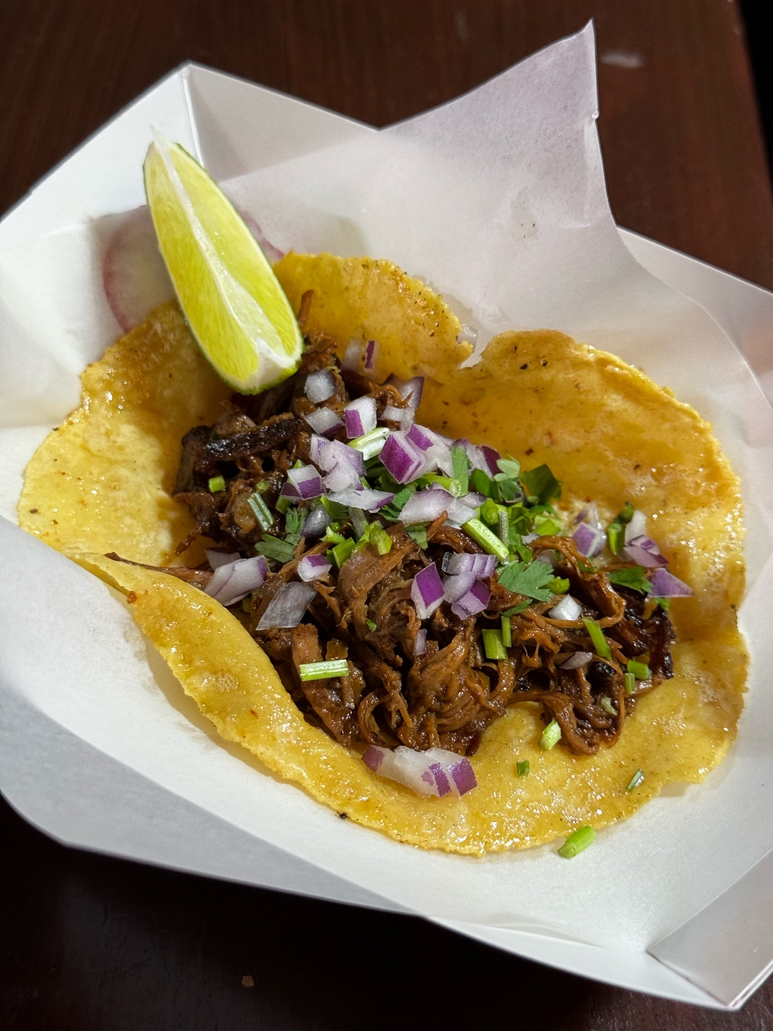 A corn tortilla topped with shredded meat, chopped red onions, green herbs, and served with a lime wedge in a paper tray.