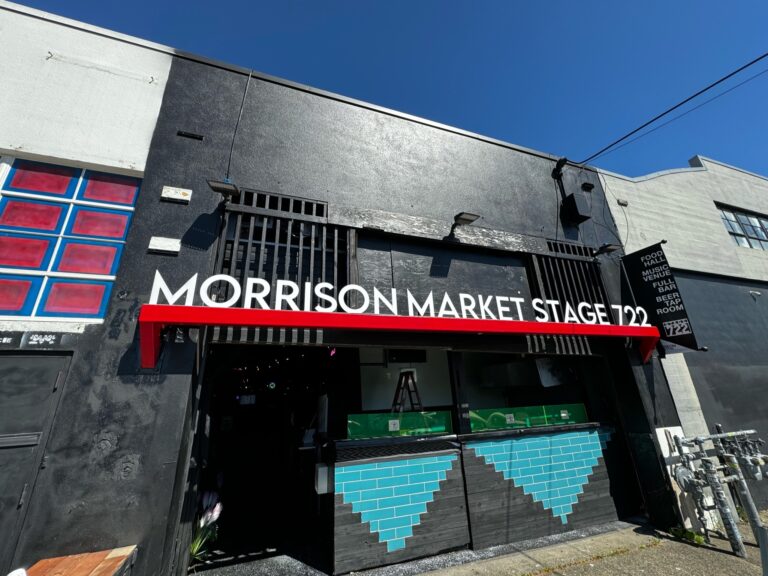 Storefront of "Morrison Market Stage 1220" with a red awning, black exterior, and blue geometric designs under the open window, set against a clear blue sky.