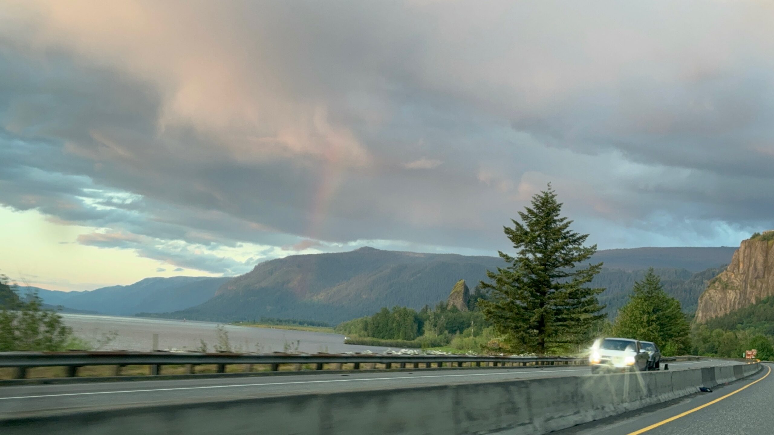 A car drives on a highway beside a large river, with mountains, trees, and a faint rainbow under a cloudy sky in the background.