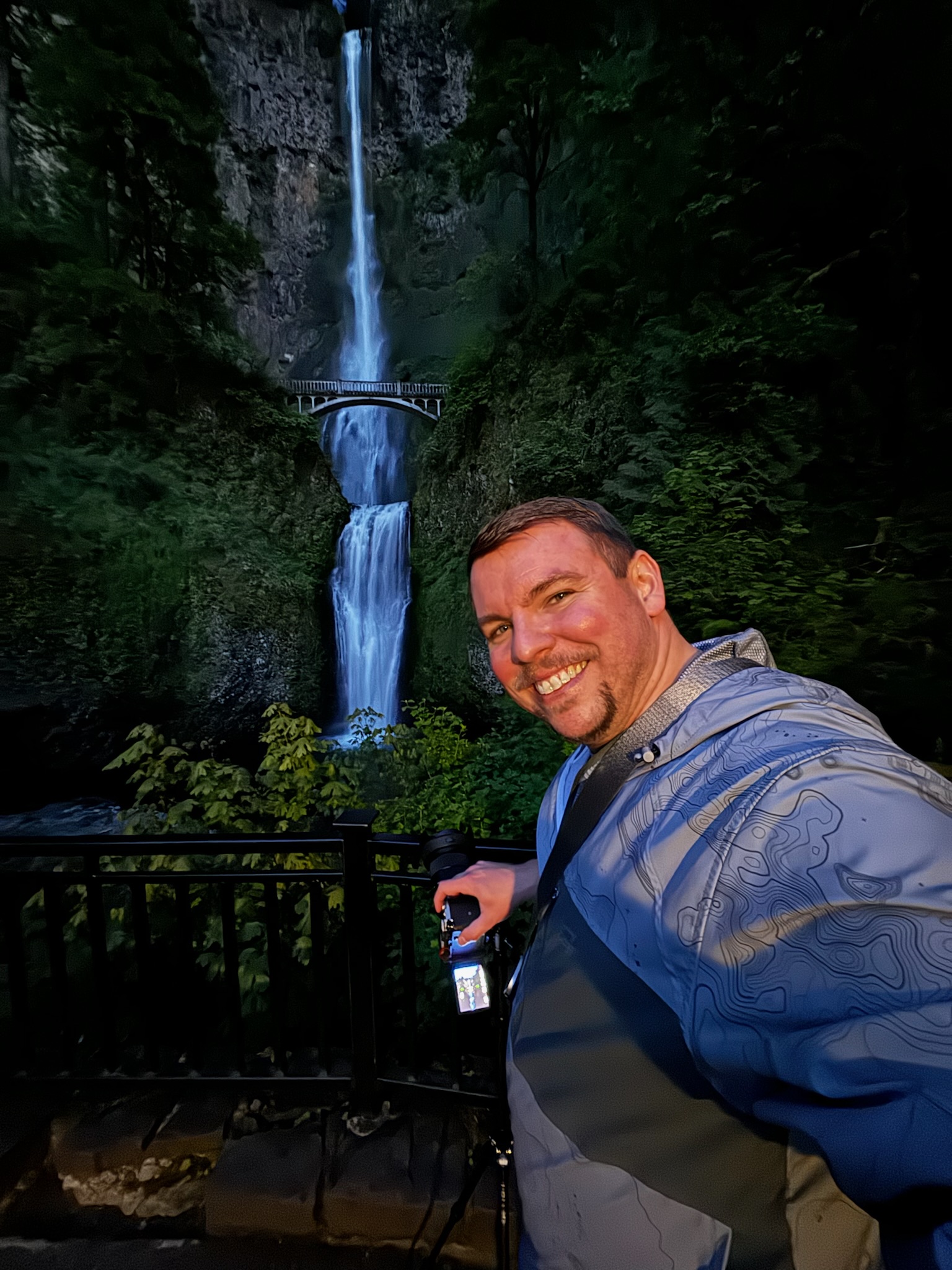 A man in a blue jacket smiles at the camera while standing in front of a waterfall and a footbridge, surrounded by lush greenery at dusk.