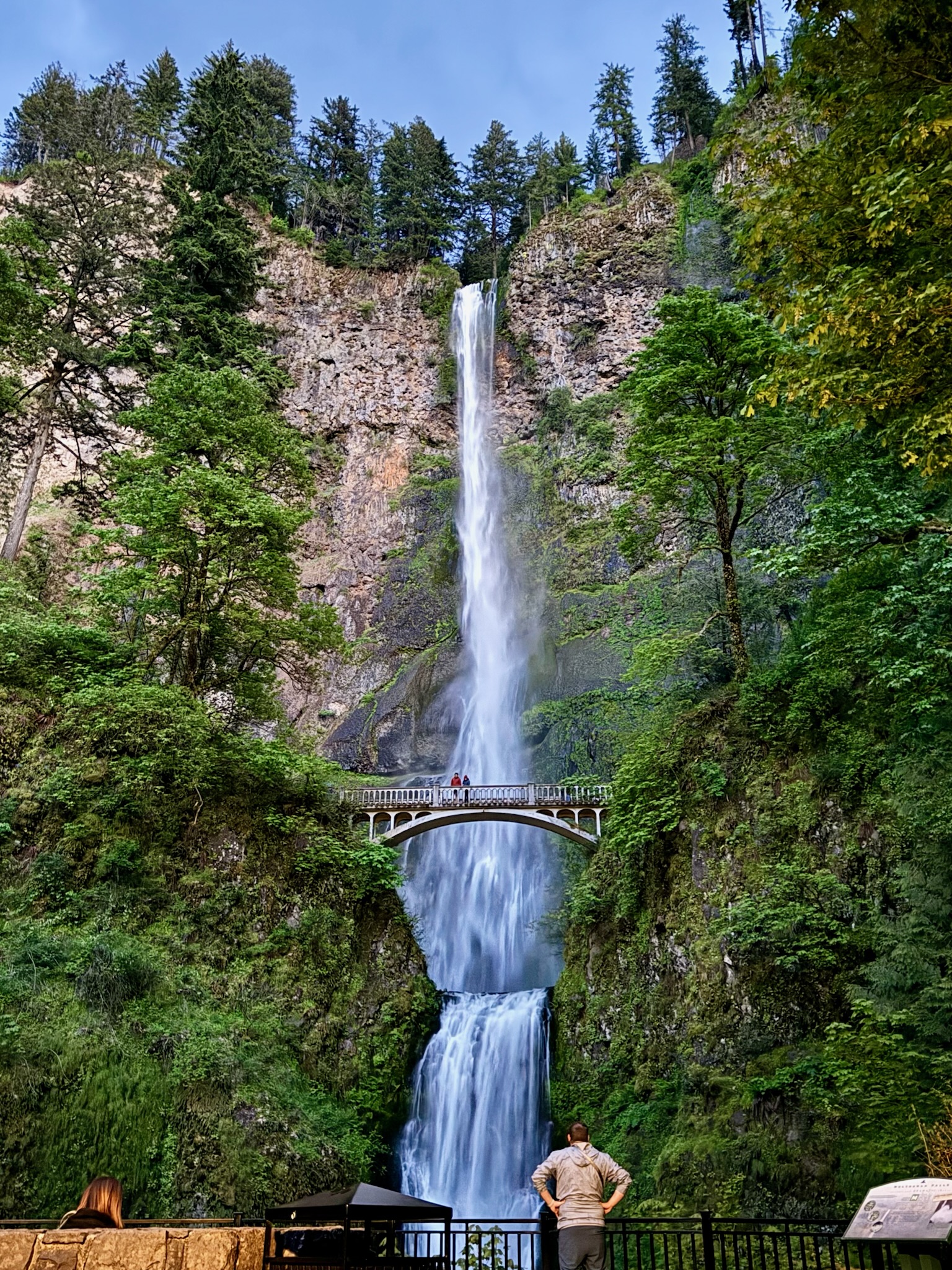 A tall waterfall cascades down a rocky cliff surrounded by trees, with a footbridge spanning the lower falls and people observing from below.