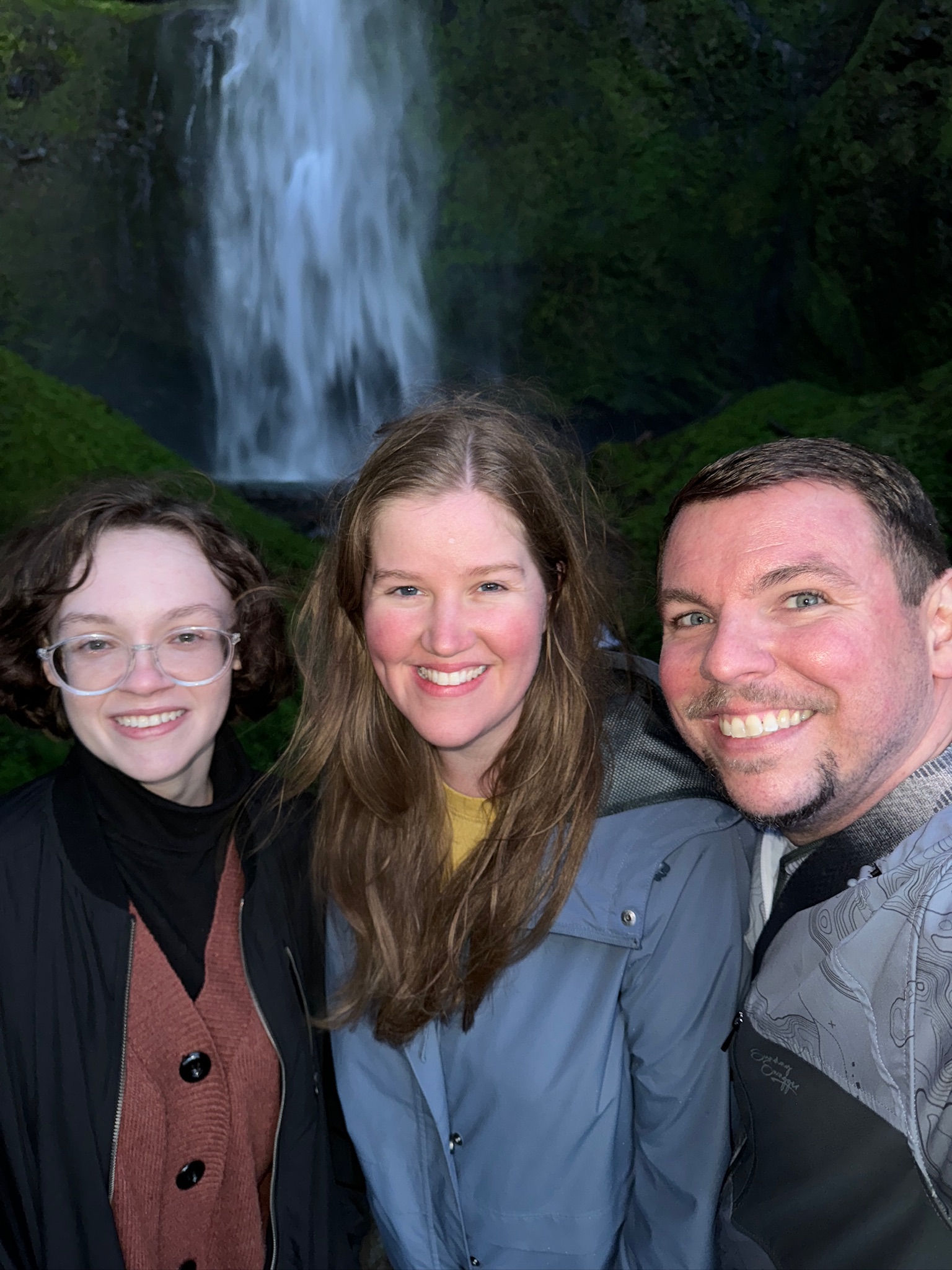 Three people smiling and posing for a photo in front of a waterfall with green mossy surroundings.