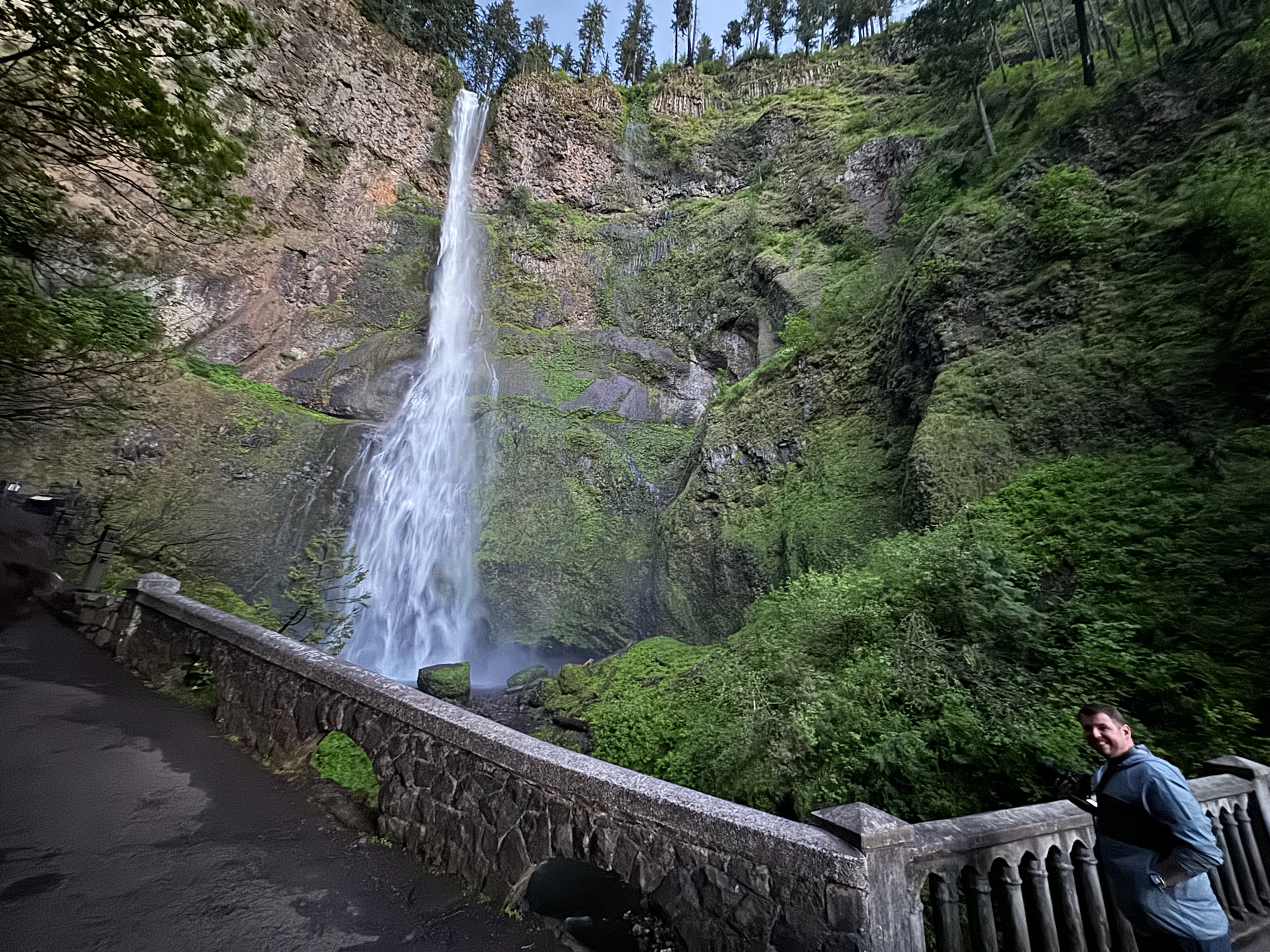 A tall waterfall cascades down a steep, rocky, green-covered cliff near a stone bridge, with a person standing and looking at the camera in the foreground.