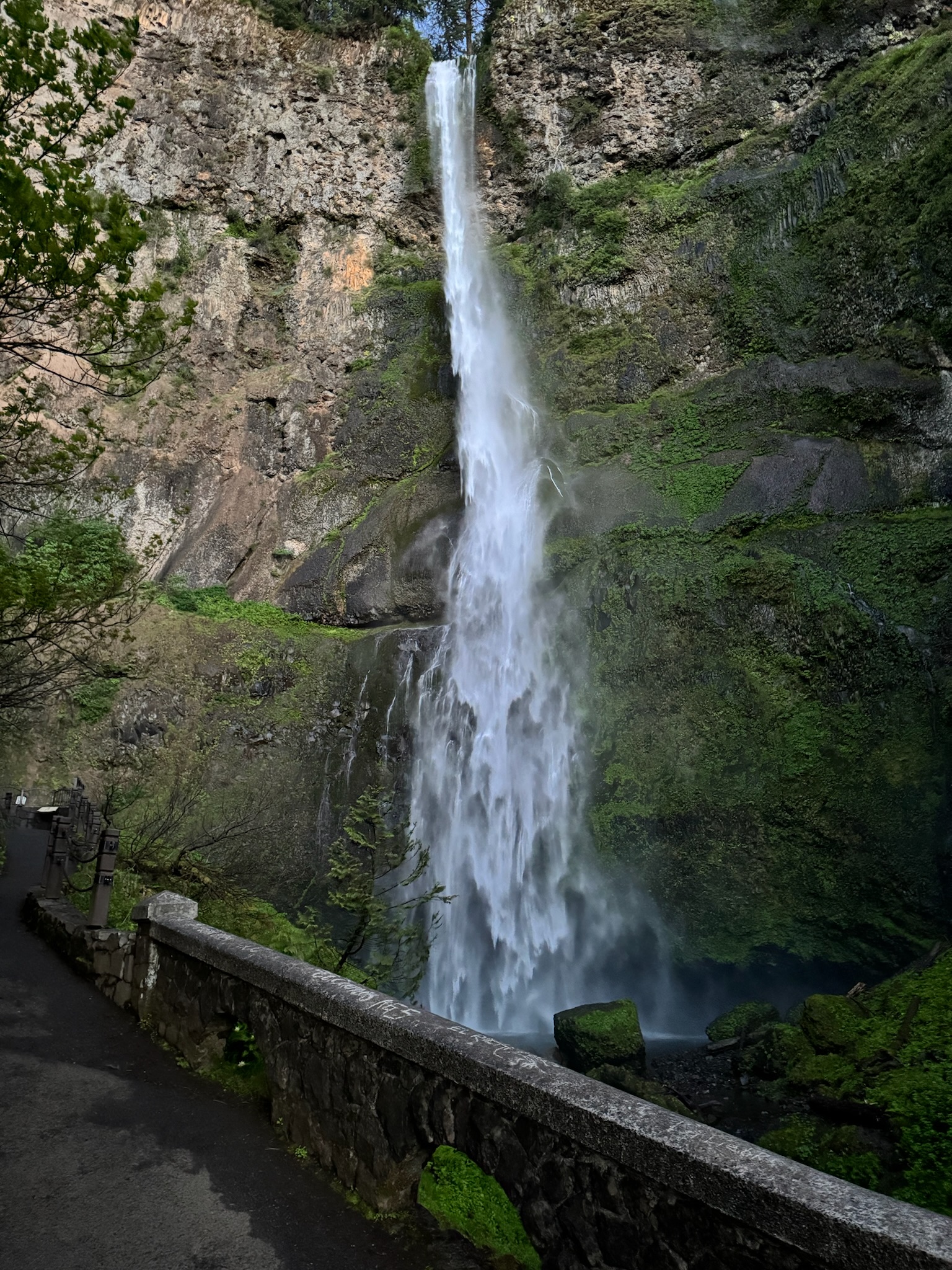 A tall waterfall cascades down a rocky, moss-covered cliff surrounded by greenery, with a stone railing and pathway in the foreground.