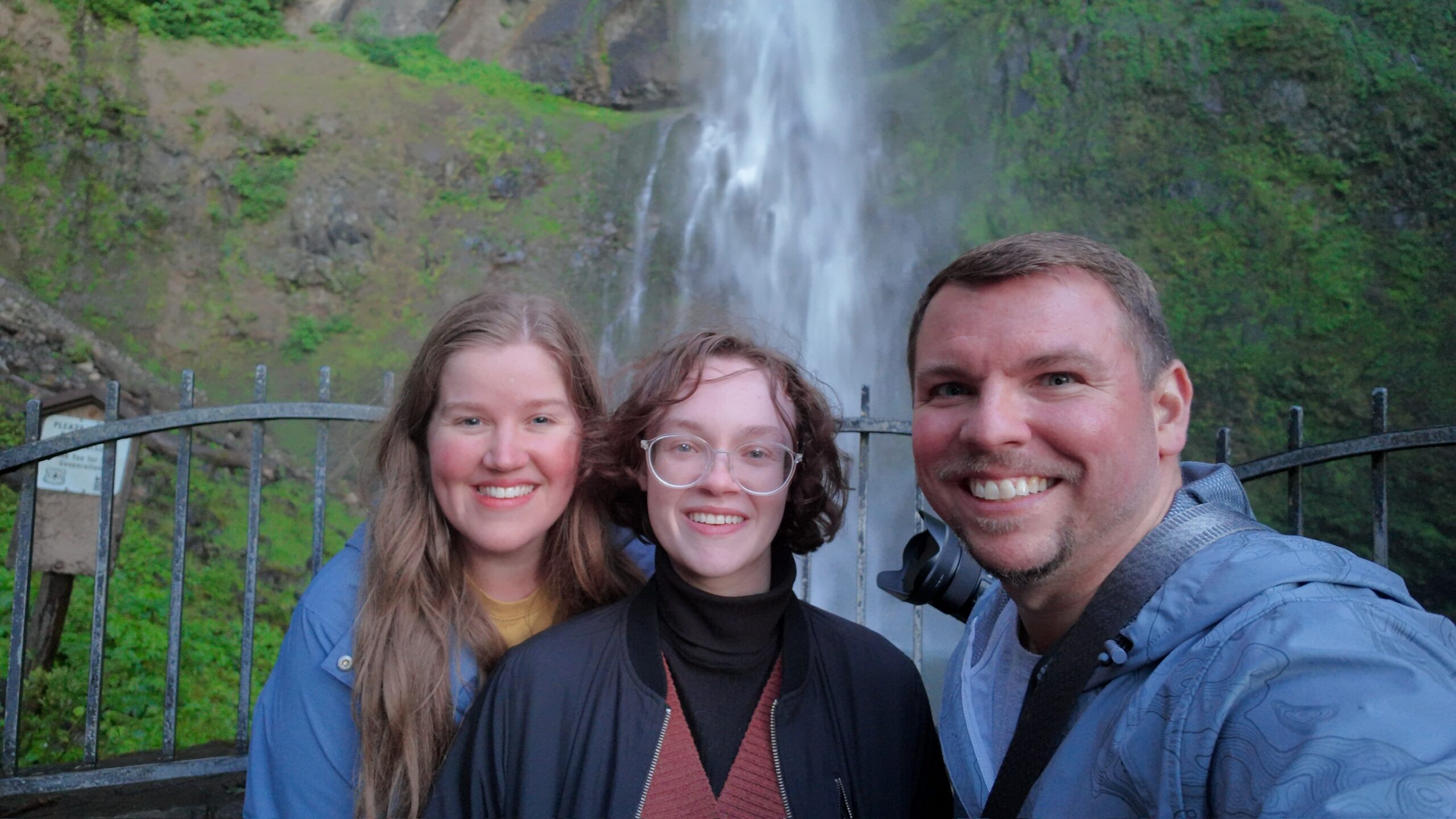 Three people smiling in front of a metal fence with a waterfall and greenery in the background.