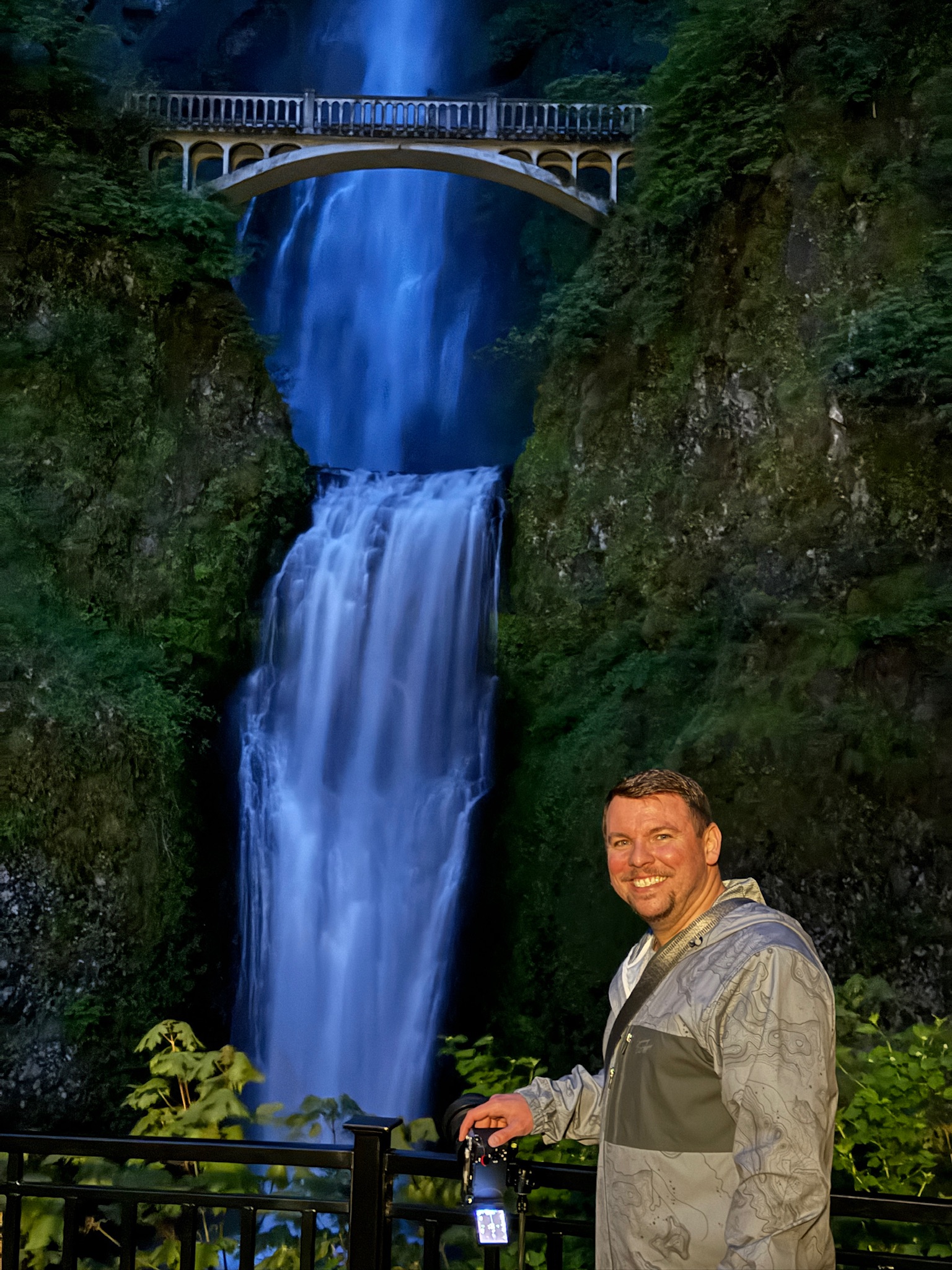 A man in a gray jacket stands smiling in front of Multnomah Falls with a stone bridge above the waterfall, surrounded by greenery.