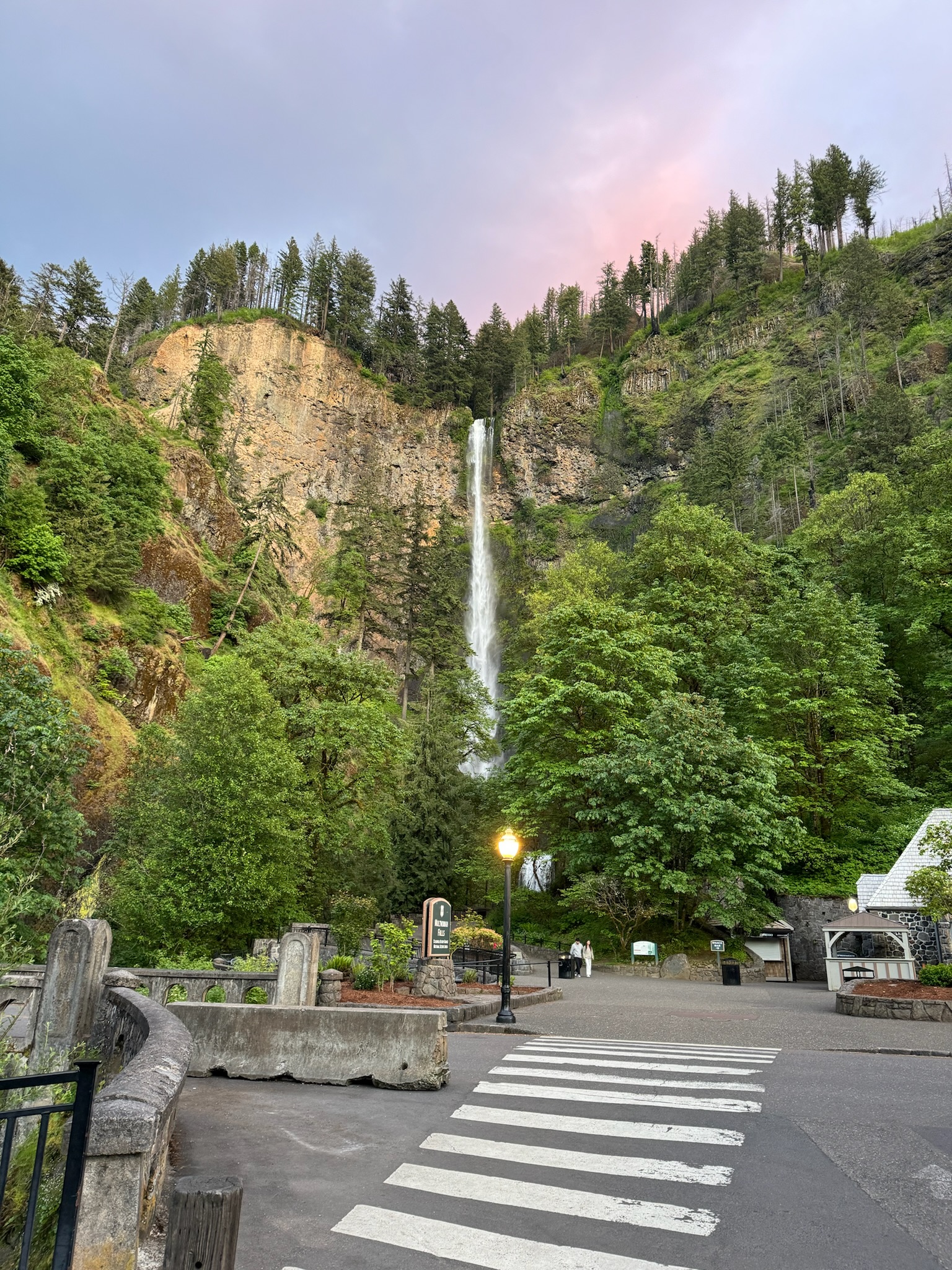 A tall waterfall cascades down a rocky cliff surrounded by dense green trees, with a crosswalk and lamppost in the foreground under a partly cloudy sky.