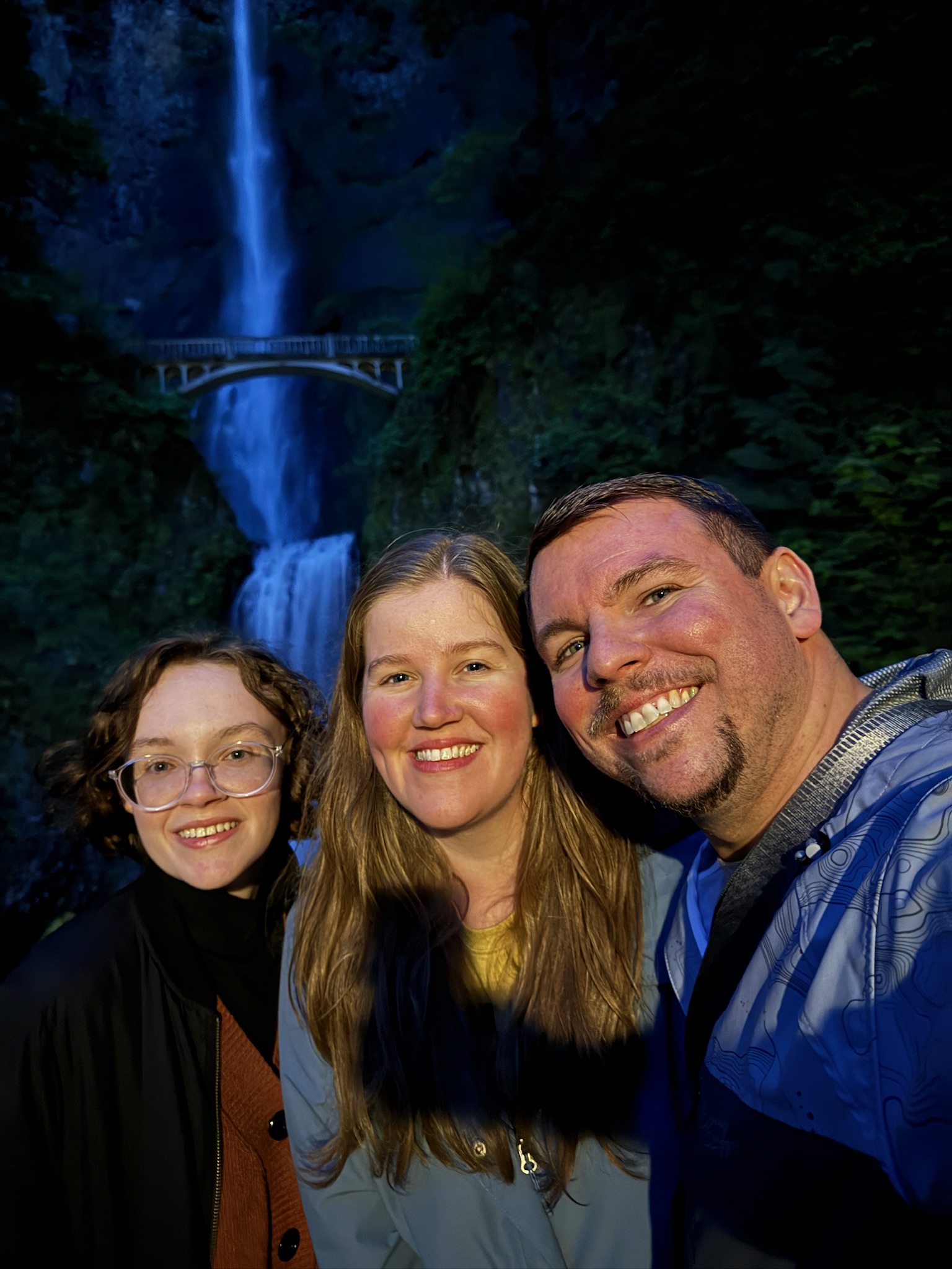 Three people smile for a selfie in front of a tall waterfall and footbridge surrounded by lush greenery at dusk.