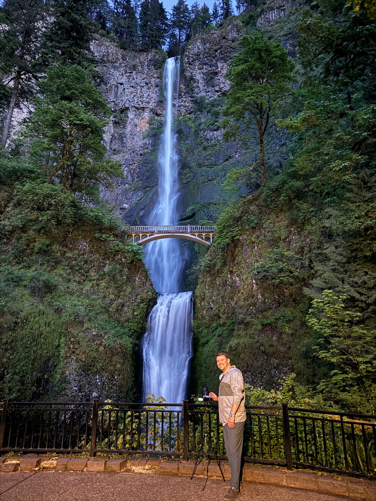 A man stands next to a railing with a camera and tripod in front of a tall waterfall cascading down a rocky cliff, with a footbridge spanning the falls.