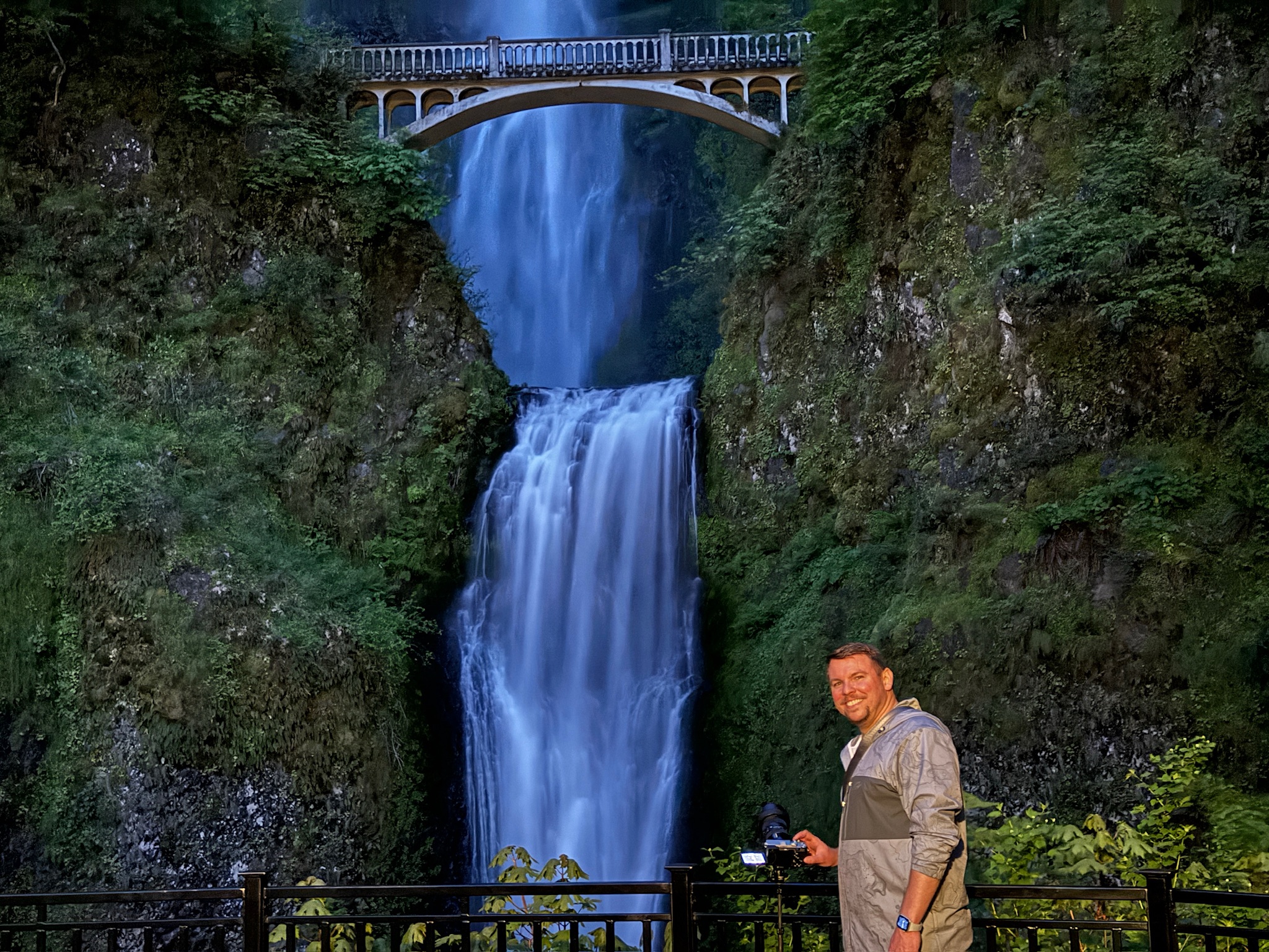 A man stands in front of a railing with a camera in hand, smiling, with Multnomah Falls and a bridge visible in the background.