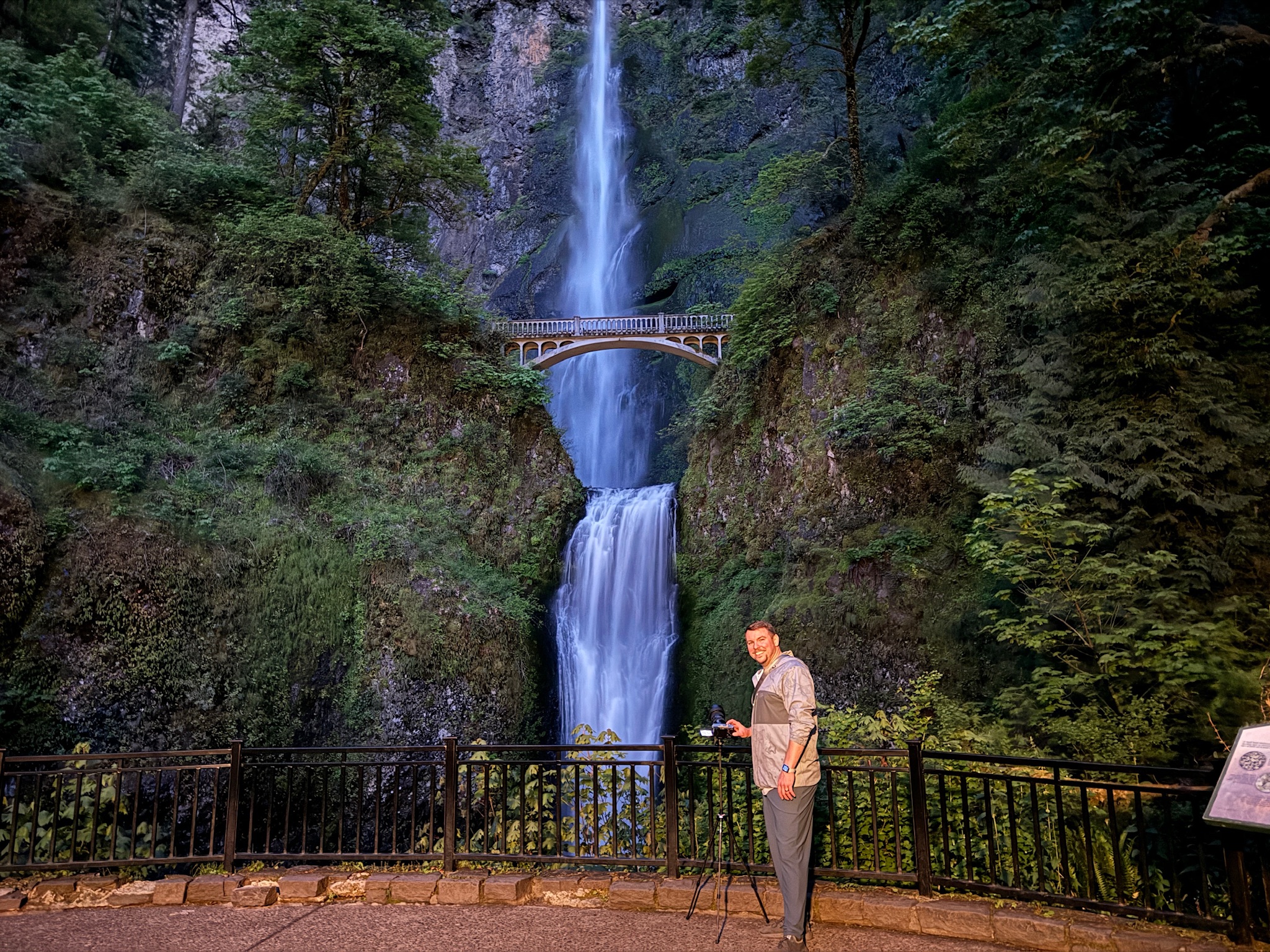A man stands with a tripod near a railing in front of Multnomah Falls, with a bridge spanning the waterfall in the background and dense greenery surrounding the scene.
