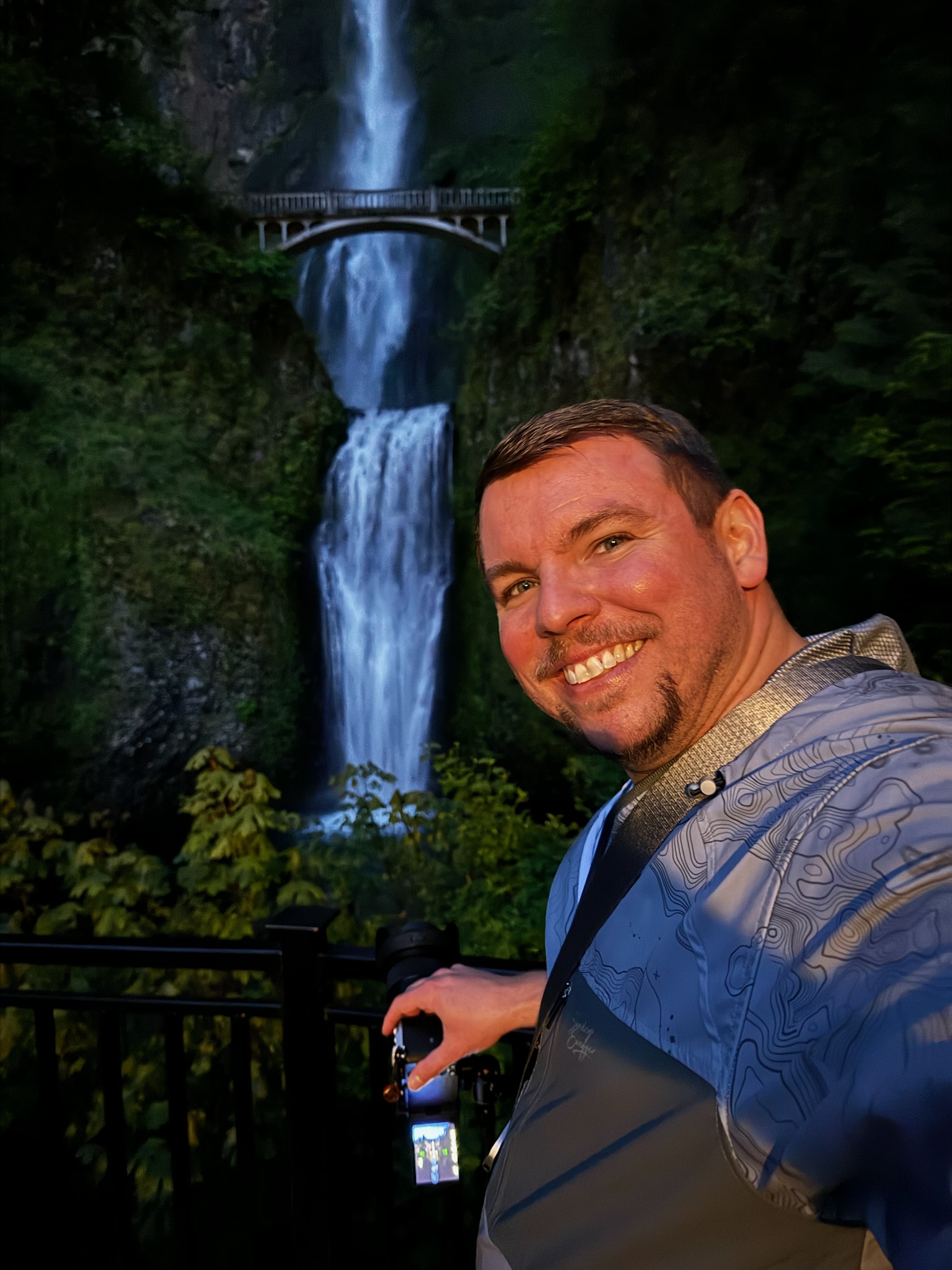A man smiles for a selfie in front of a tall, two-tiered waterfall and a bridge, holding a camera and smartphone, with greenery surrounding the scene.