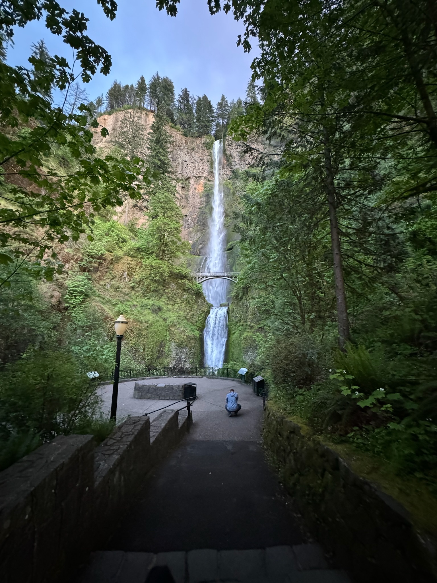 A tall waterfall cascades down a rocky cliff surrounded by lush greenery, with a stone pathway and a person sitting near the base.