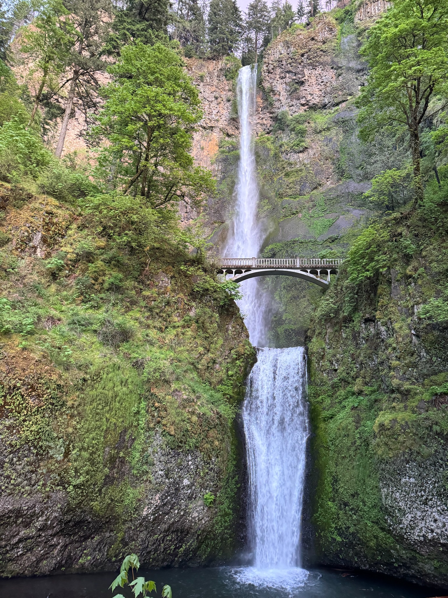 A tall waterfall cascades down a rocky cliff in two tiers, with a footbridge crossing between the upper and lower falls, surrounded by lush green trees.