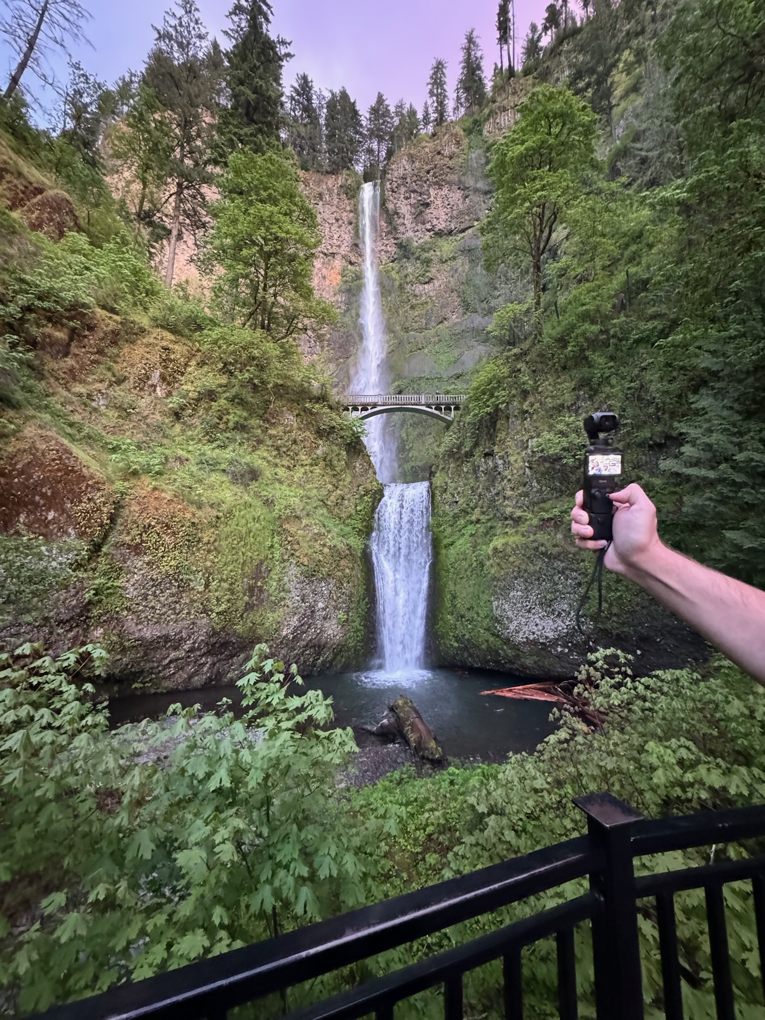 A person holds a small camera facing a two-tiered waterfall surrounded by lush greenery, with a footbridge spanning the upper cascade.