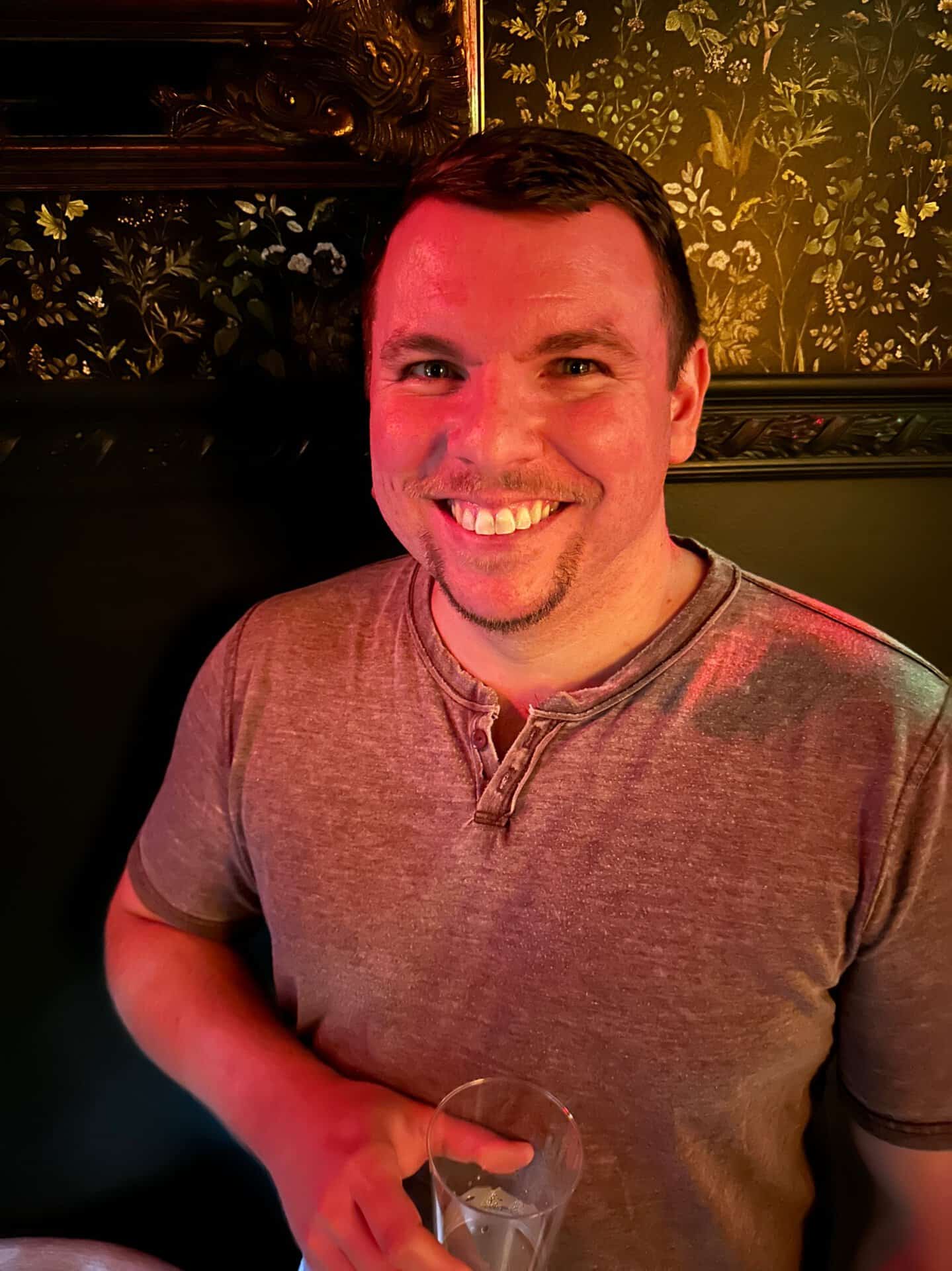 Smiling man in a grey shirt holds a glass, standing against a dark floral wallpapered background in a chic Nashville venue.