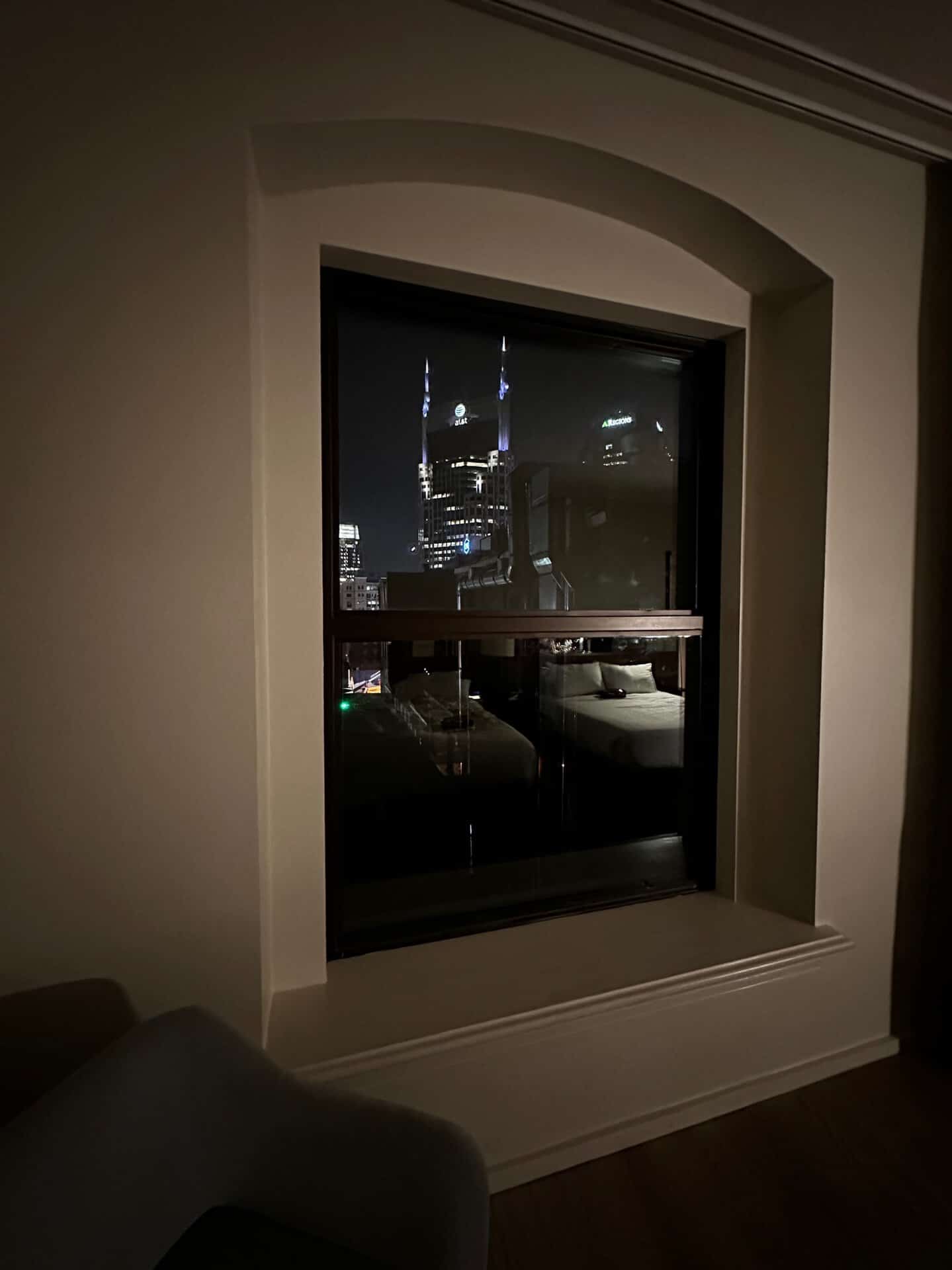 Night view through a window reveals a dimly lit room with a chair, while Nashville's bright city lights create an inviting glow outside.