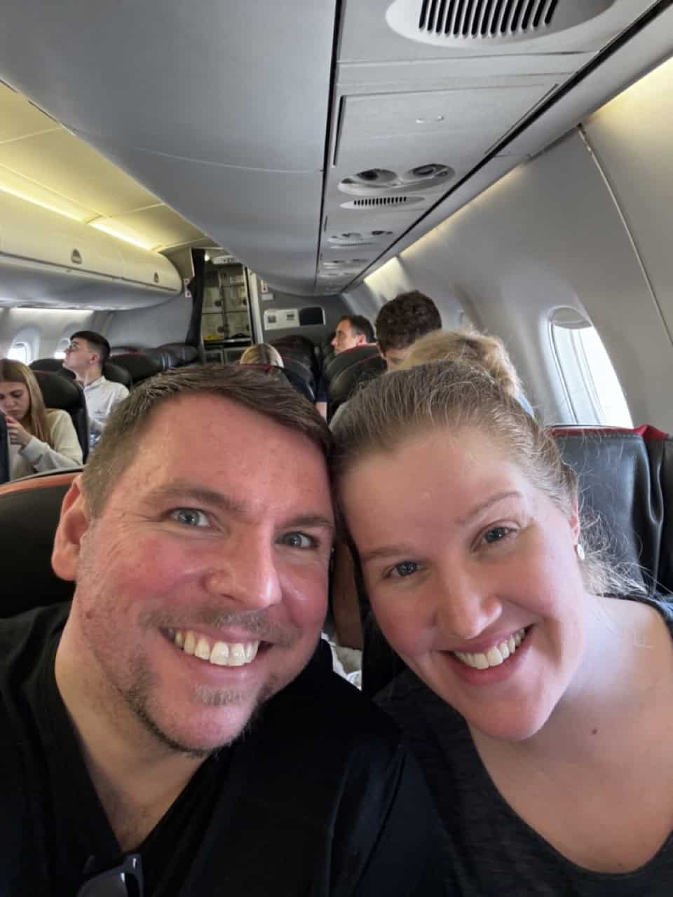 A man and a woman are all smiles, taking a selfie on their flight to Nashville, with passengers seated contentedly in the background.