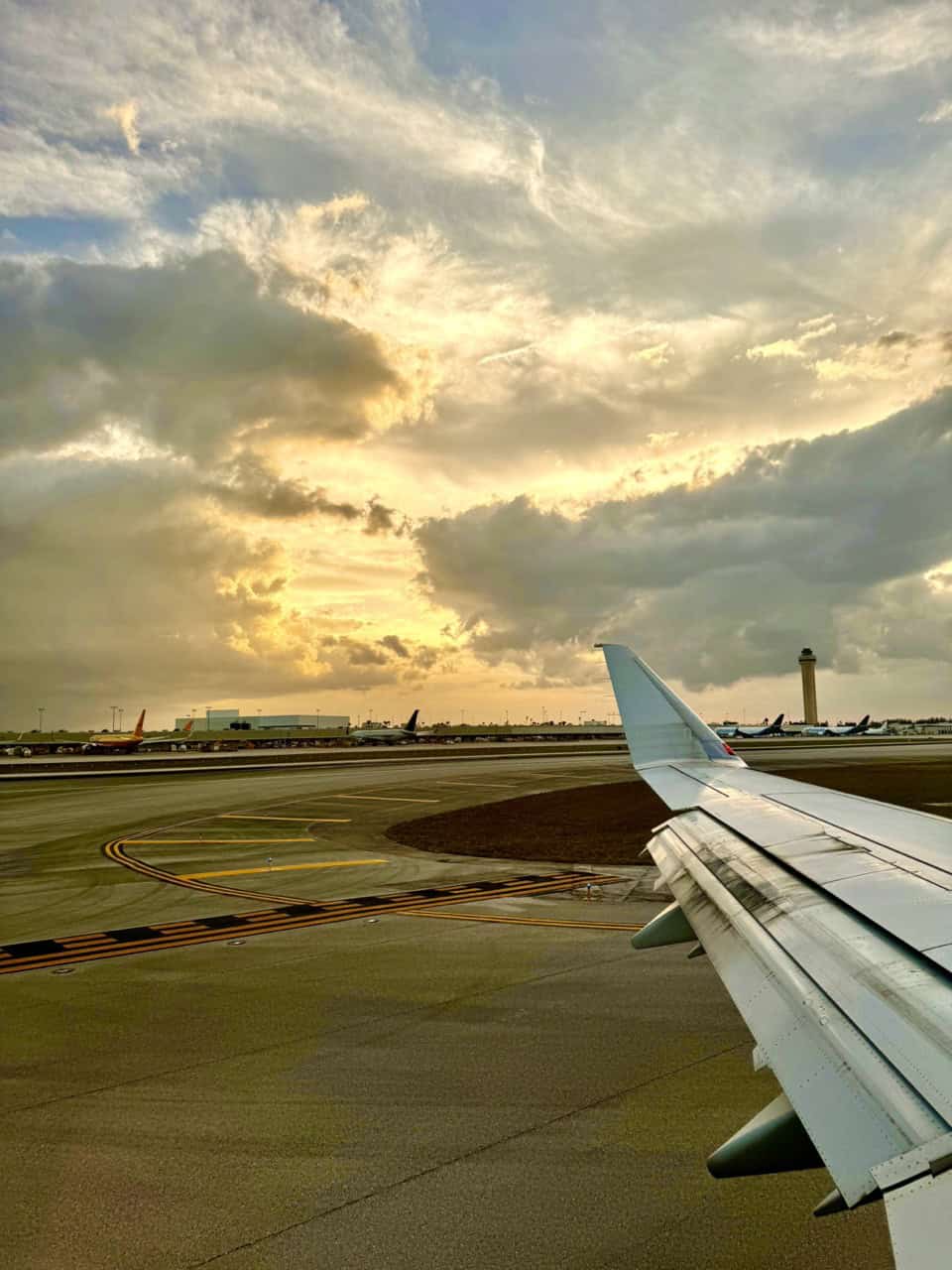 View from an airplane window as the wing glides over Nashville's airport runway. The terminal and multiple planes bask in the warm hues of a sunset against a partly cloudy sky.