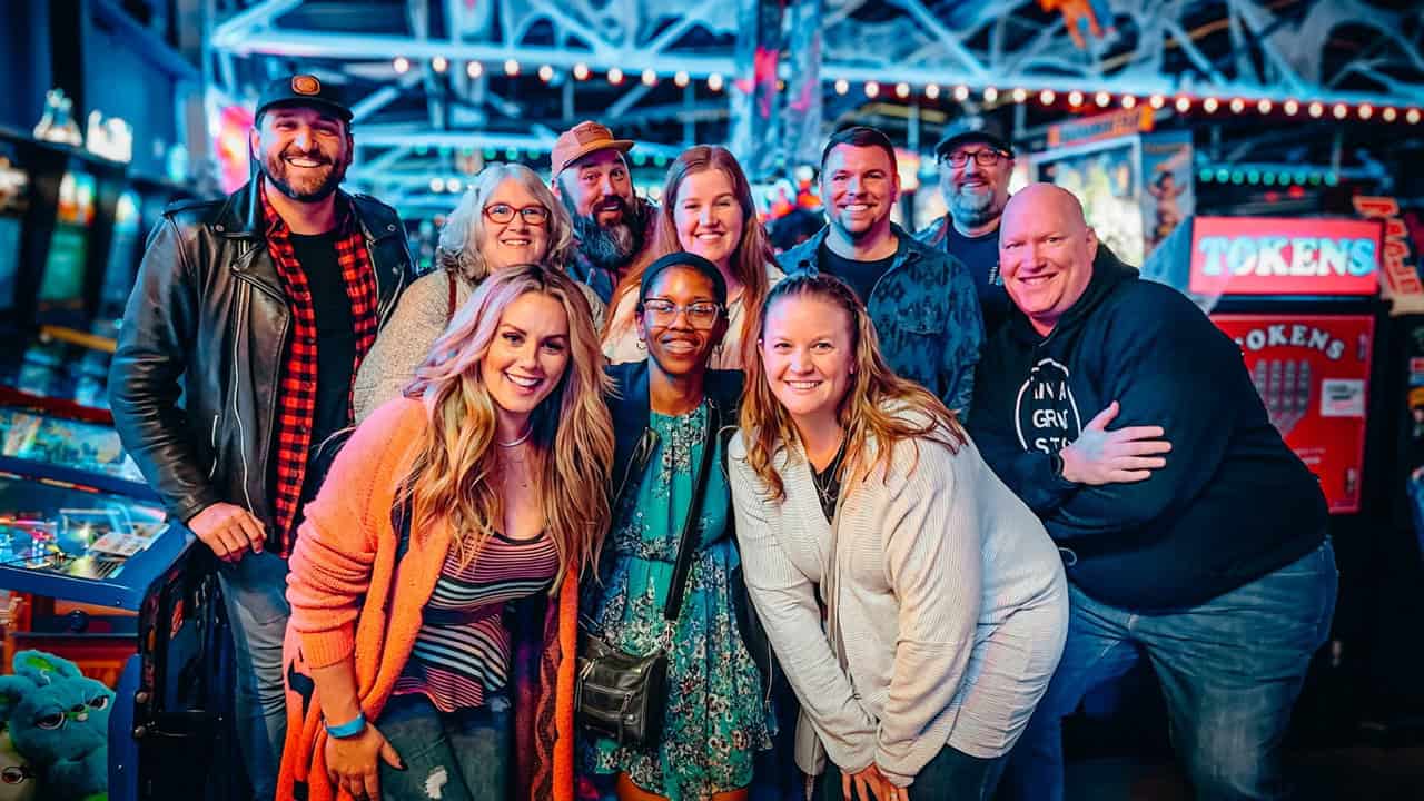 A lively group of friends is all smiles as they pose indoors, surrounded by the nostalgic glow of arcade machines and vibrant lights, capturing the spirited essence of Nashville.
