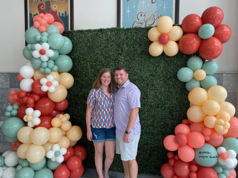 A smiling woman and man stand in front of a green leafy backdrop, surrounded by colorful balloon arrangements.
