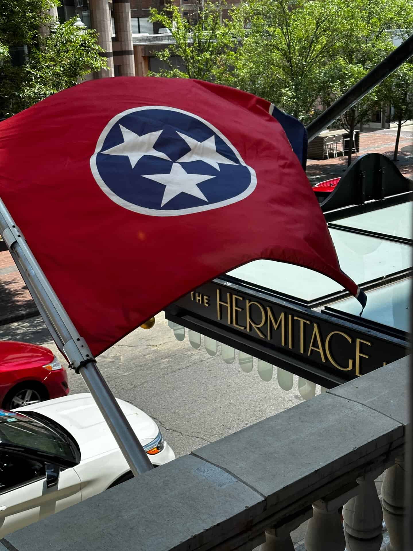 The Tennessee state flag waves proudly outside The Hermitage Hotel entrance in Nashville, with red and white cars parked on the street below.