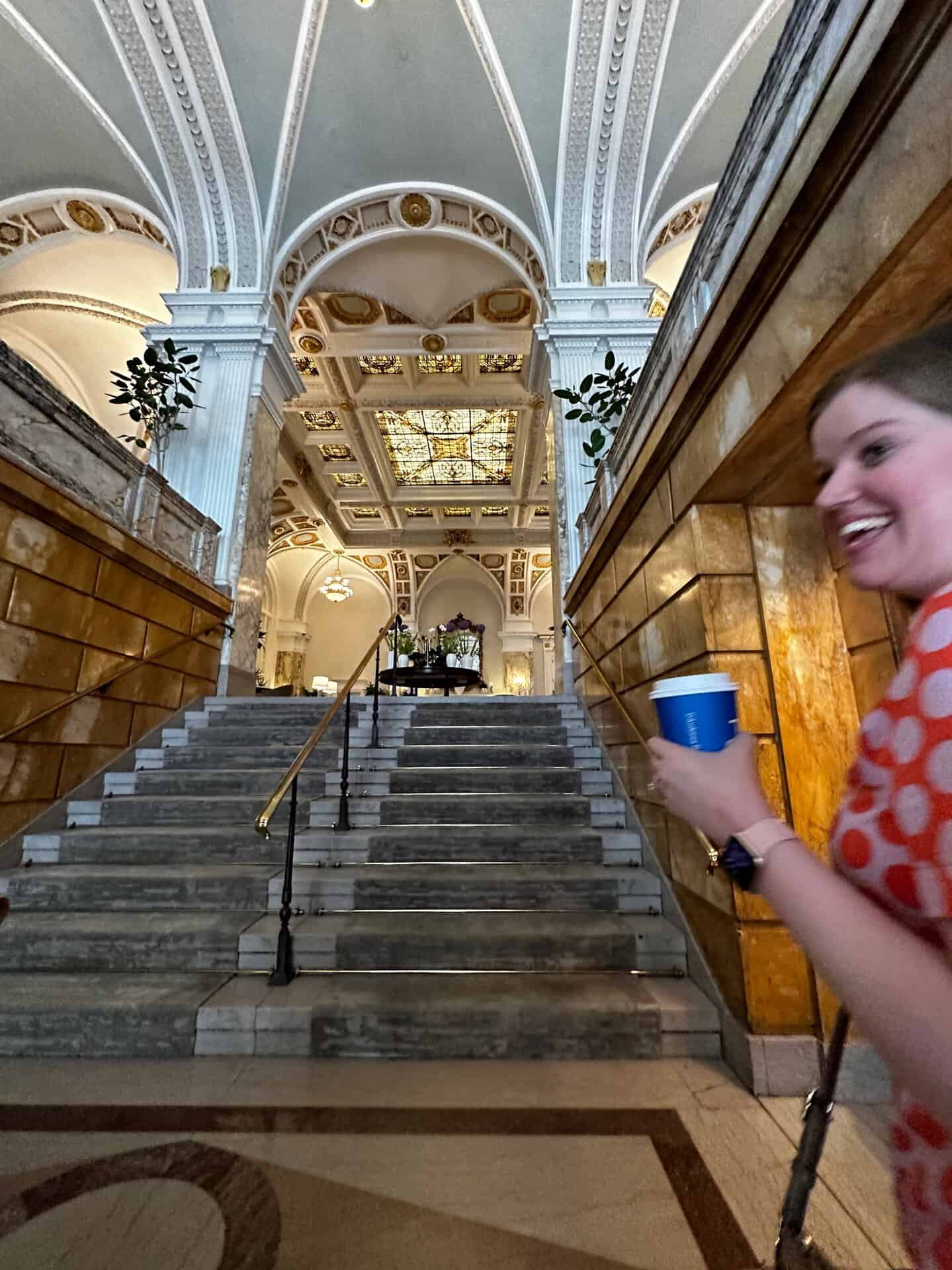 A woman holding a coffee cup strolls past a marble staircase in an ornate Nashville building, adorned with arched ceilings and stained glass.